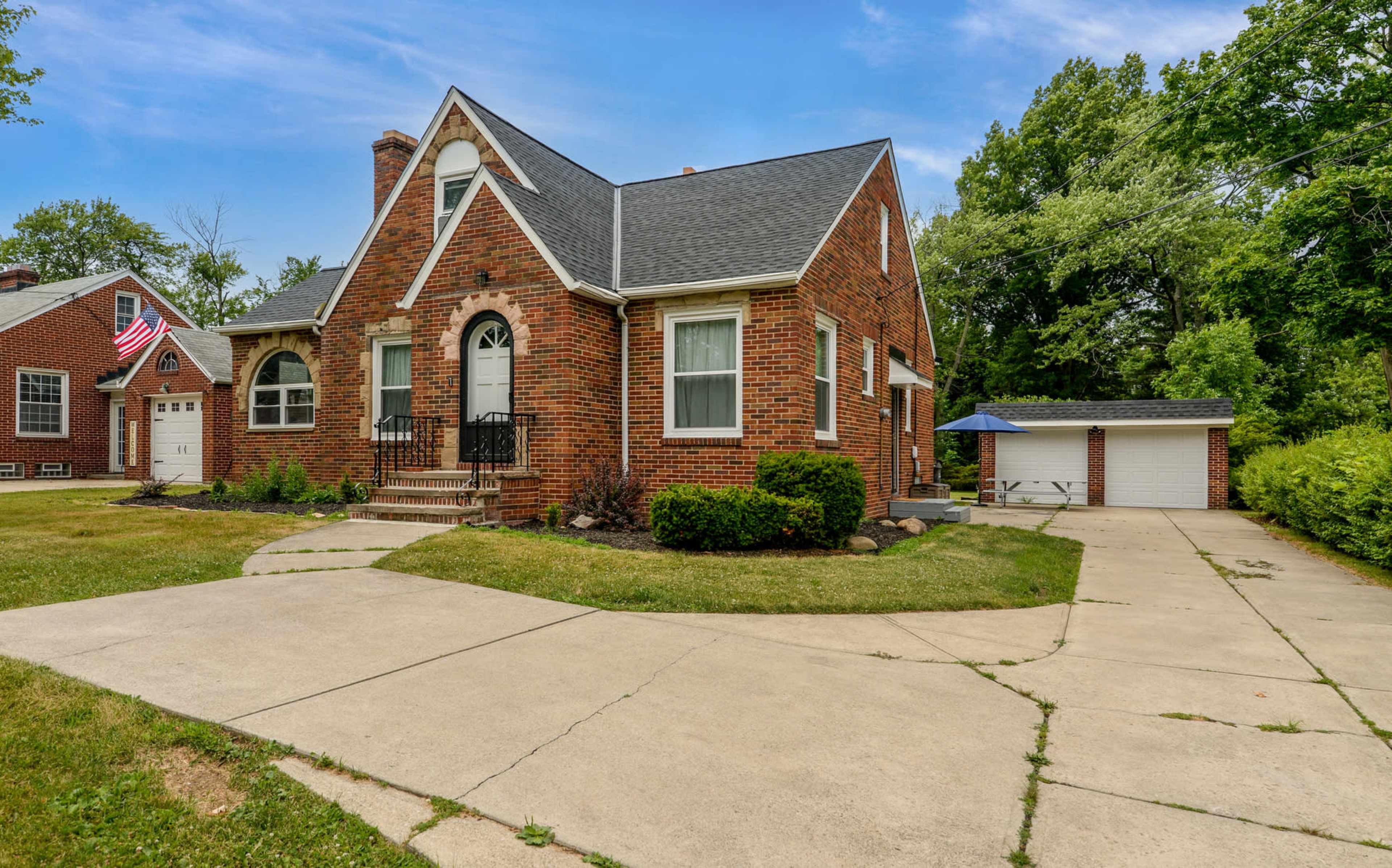 A brick house with a gabled roof is situated on a grassy lot with a concrete driveway and a garage in the background.