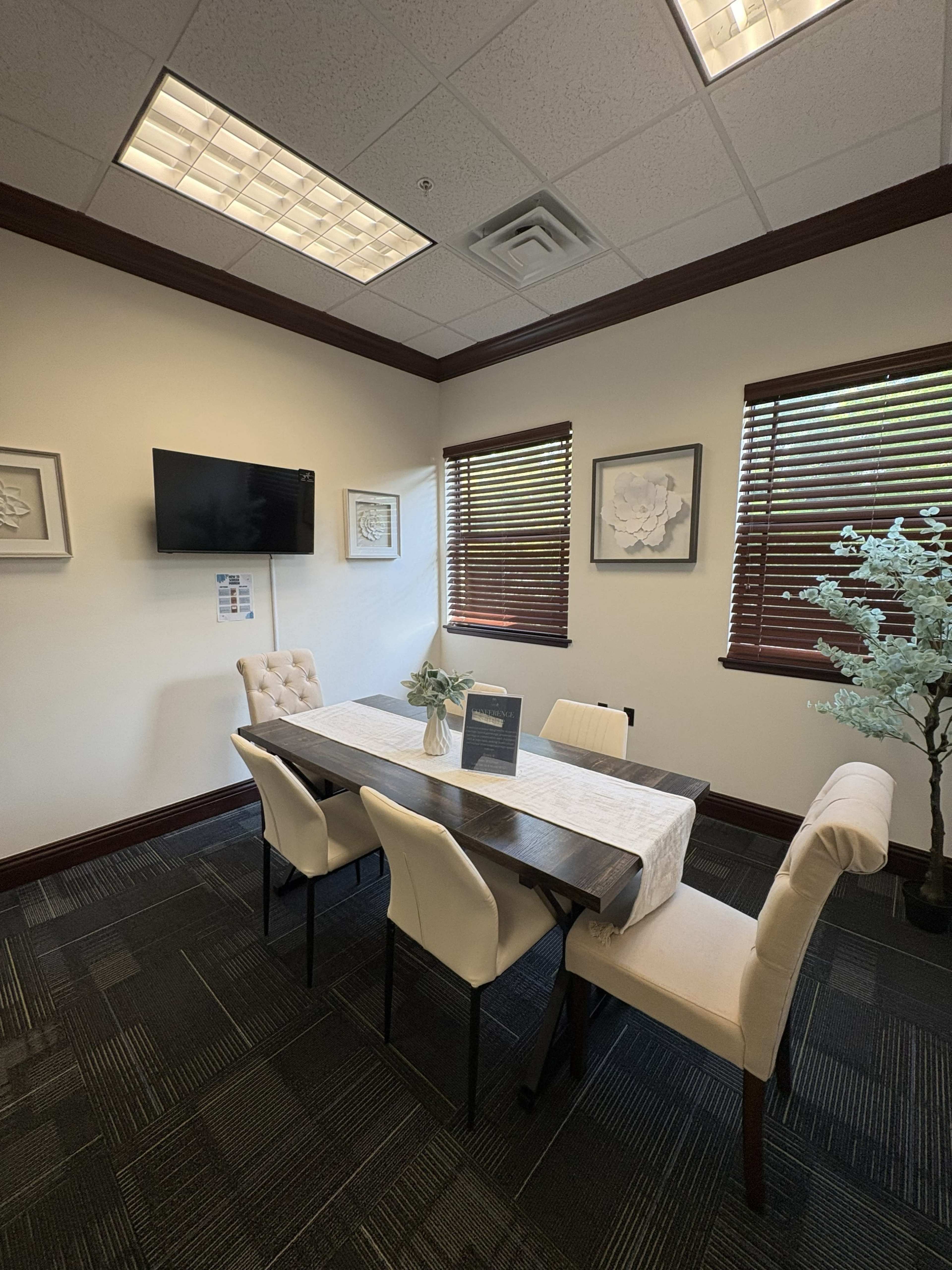 The image shows a conference room with a rectangular wooden table surrounded by four upholstered chairs and a decorative plant in the corner.