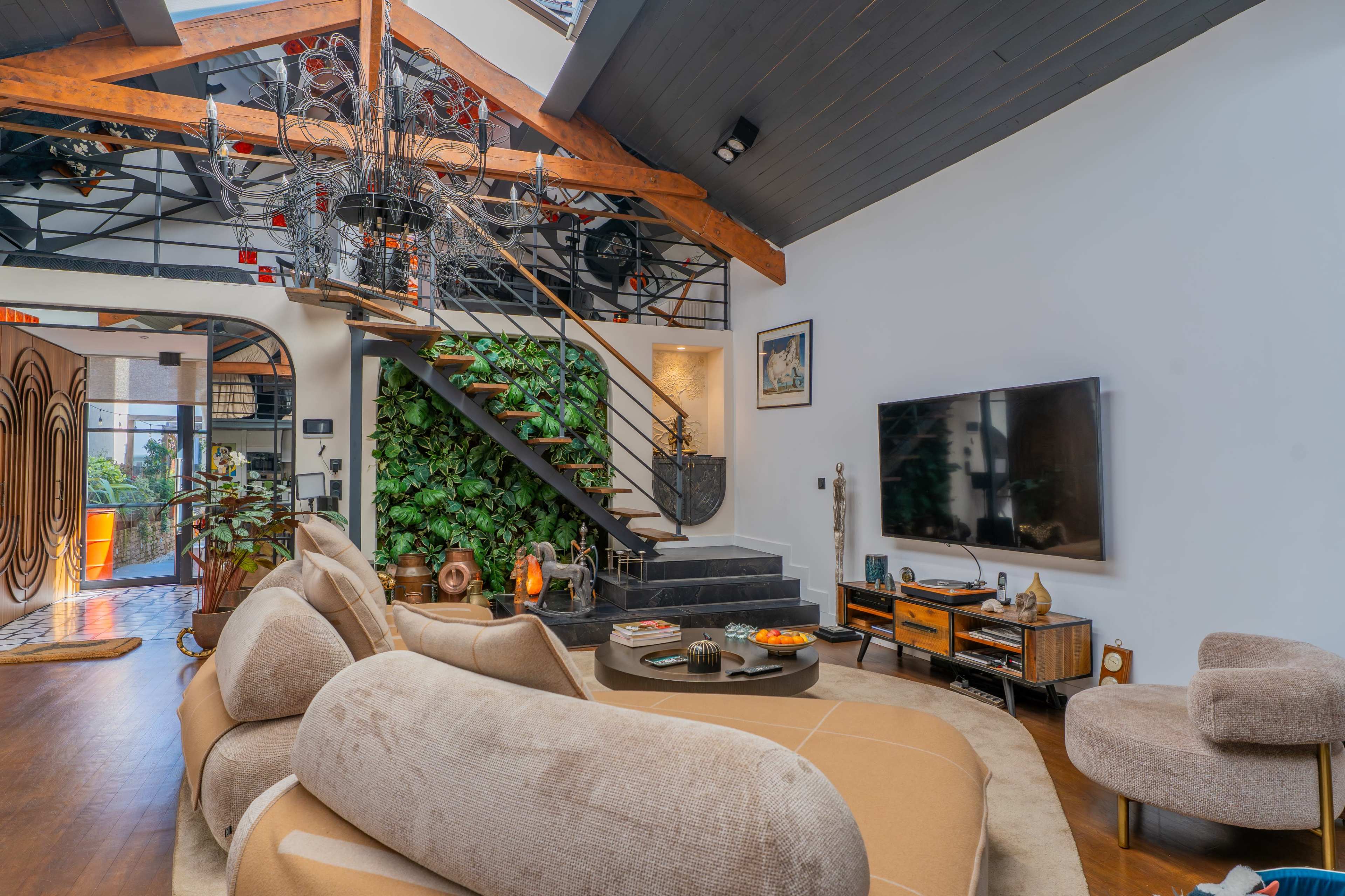 The image shows a modern living room with a curved sectional sofa, a staircase leading to an upper level, a large wall-mounted television, and decorative greenery.