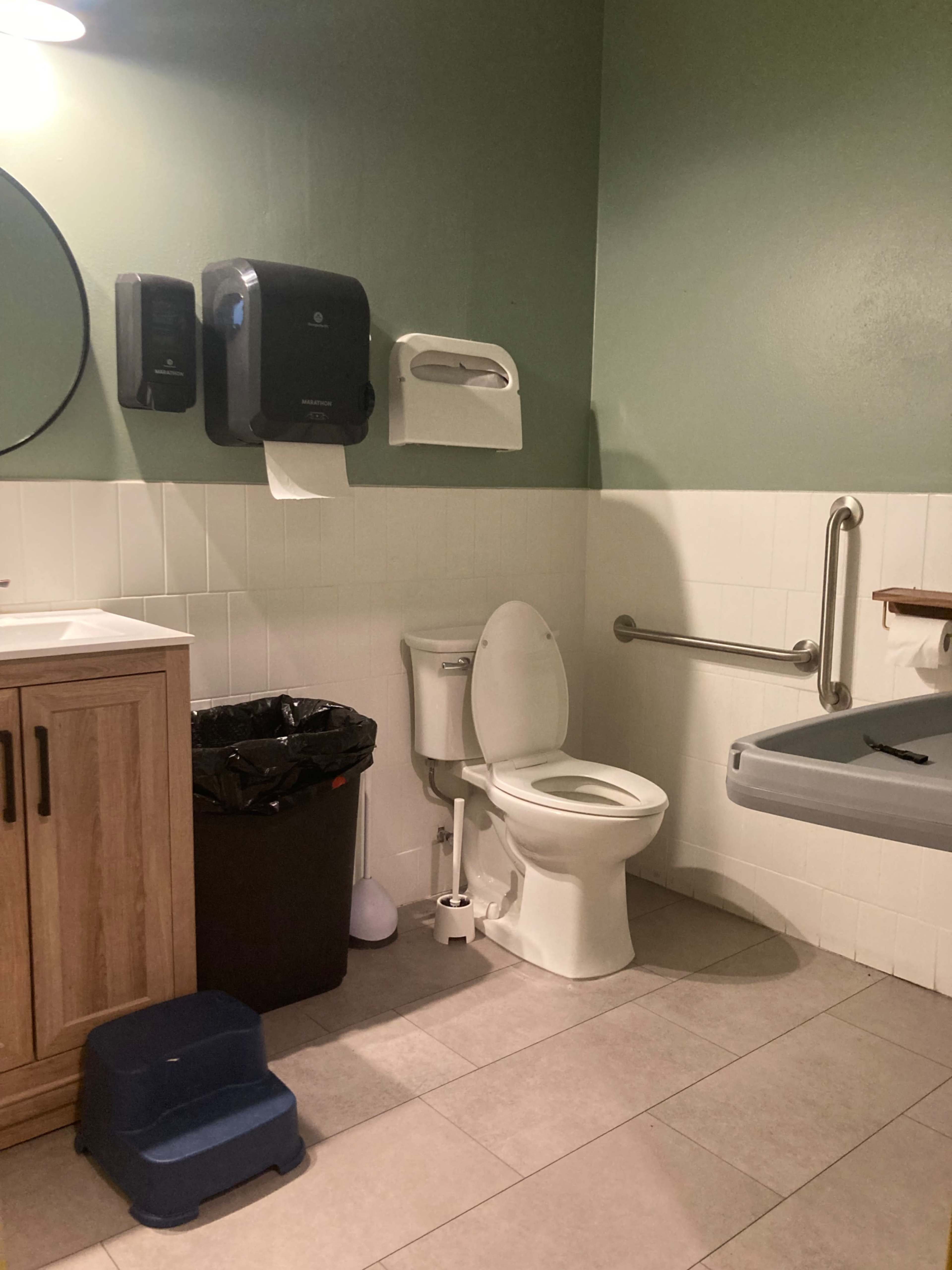 A small bathroom with a toilet, a sink, a trash can, and a changing table, along with a mirror and paper towel dispenser on the wall.