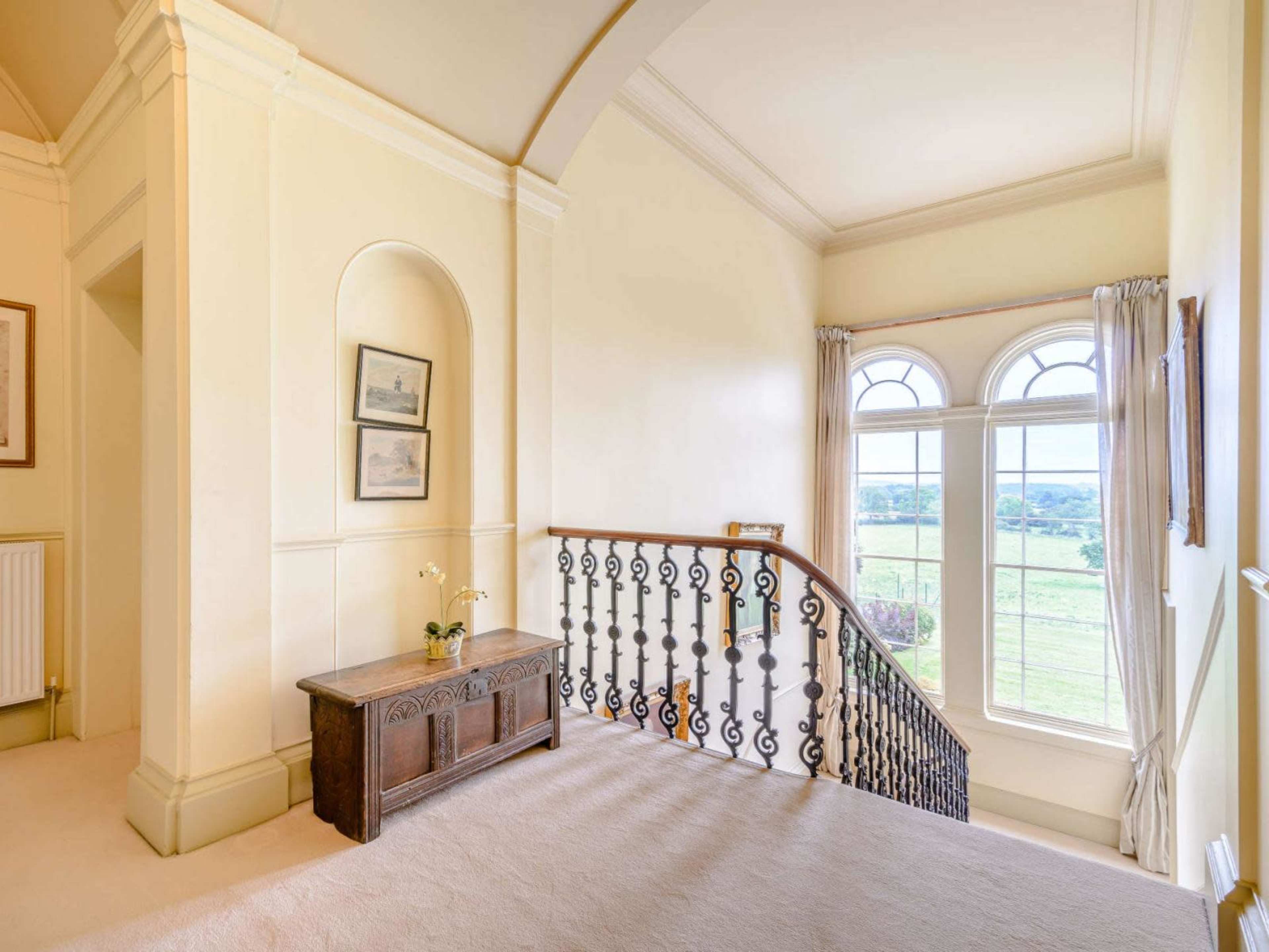 A staircase landing features a wooden railing, a small table, and large windows overlooking a grassy landscape.