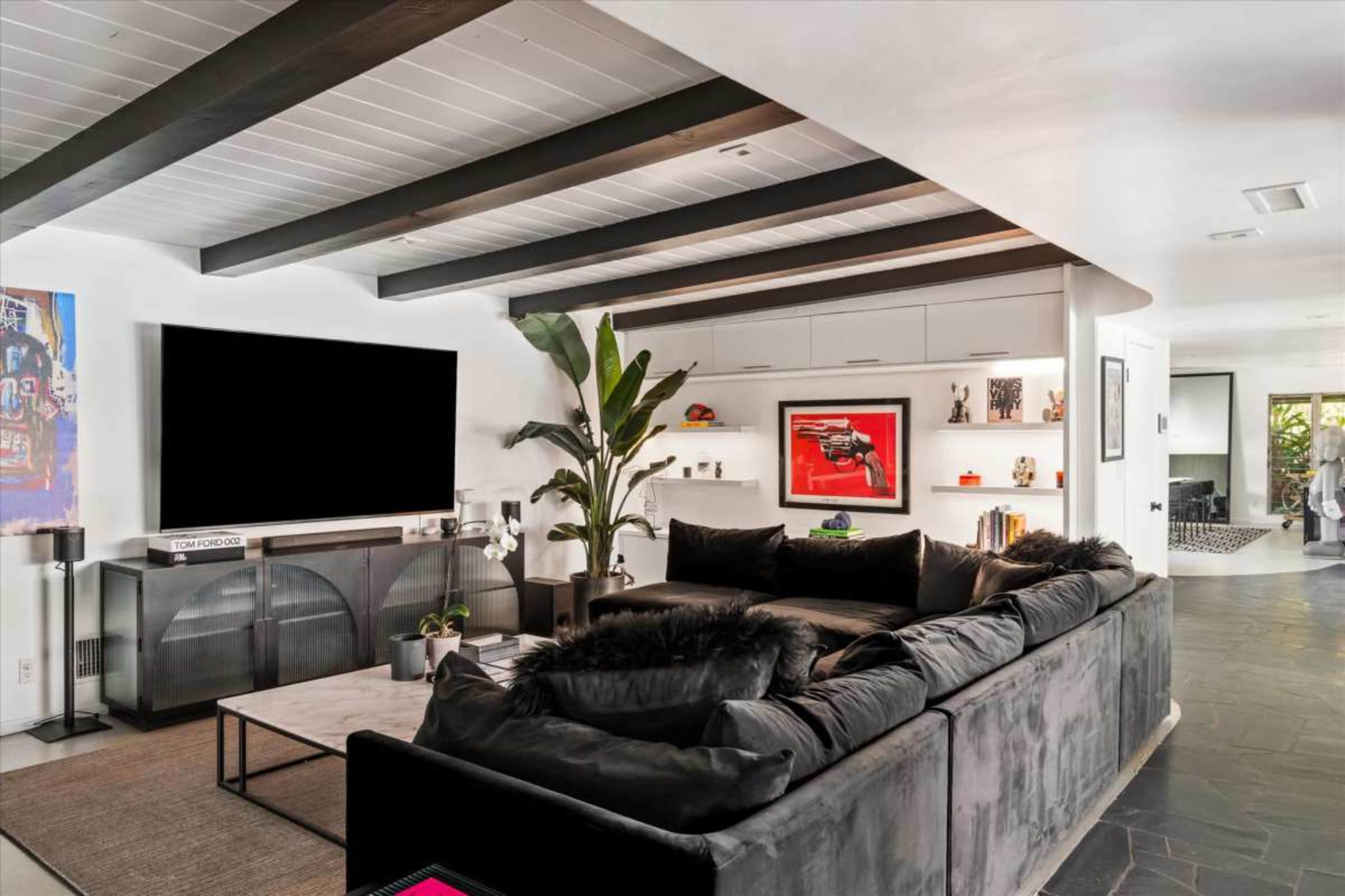 The image shows a modern living room featuring a large sectional sofa, a coffee table, and a mounted television, with a plant and artwork on the walls.