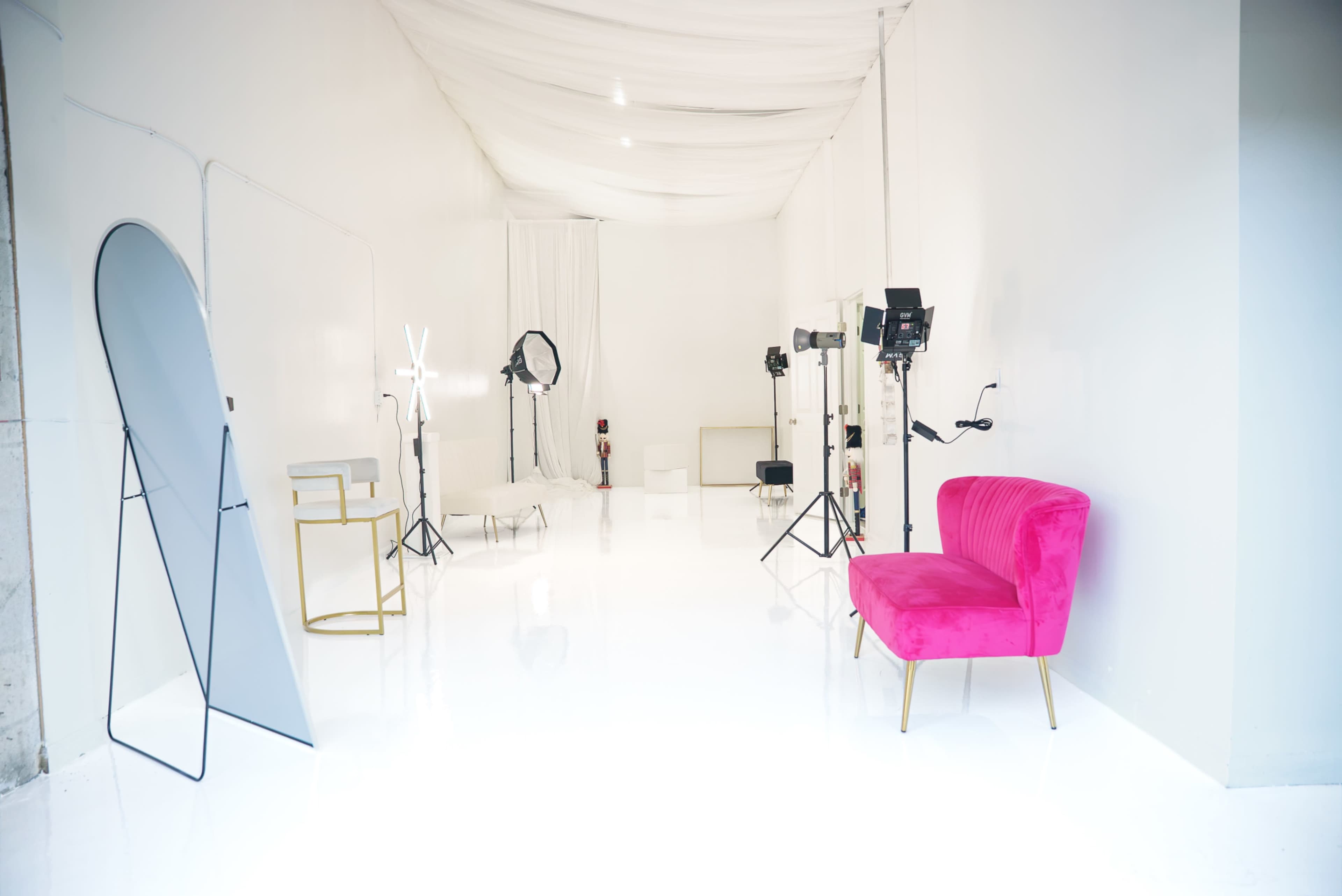 The photo shows a spacious photography studio with white walls and floor, featuring a pink velvet couch, a mirror, and various lighting equipment.