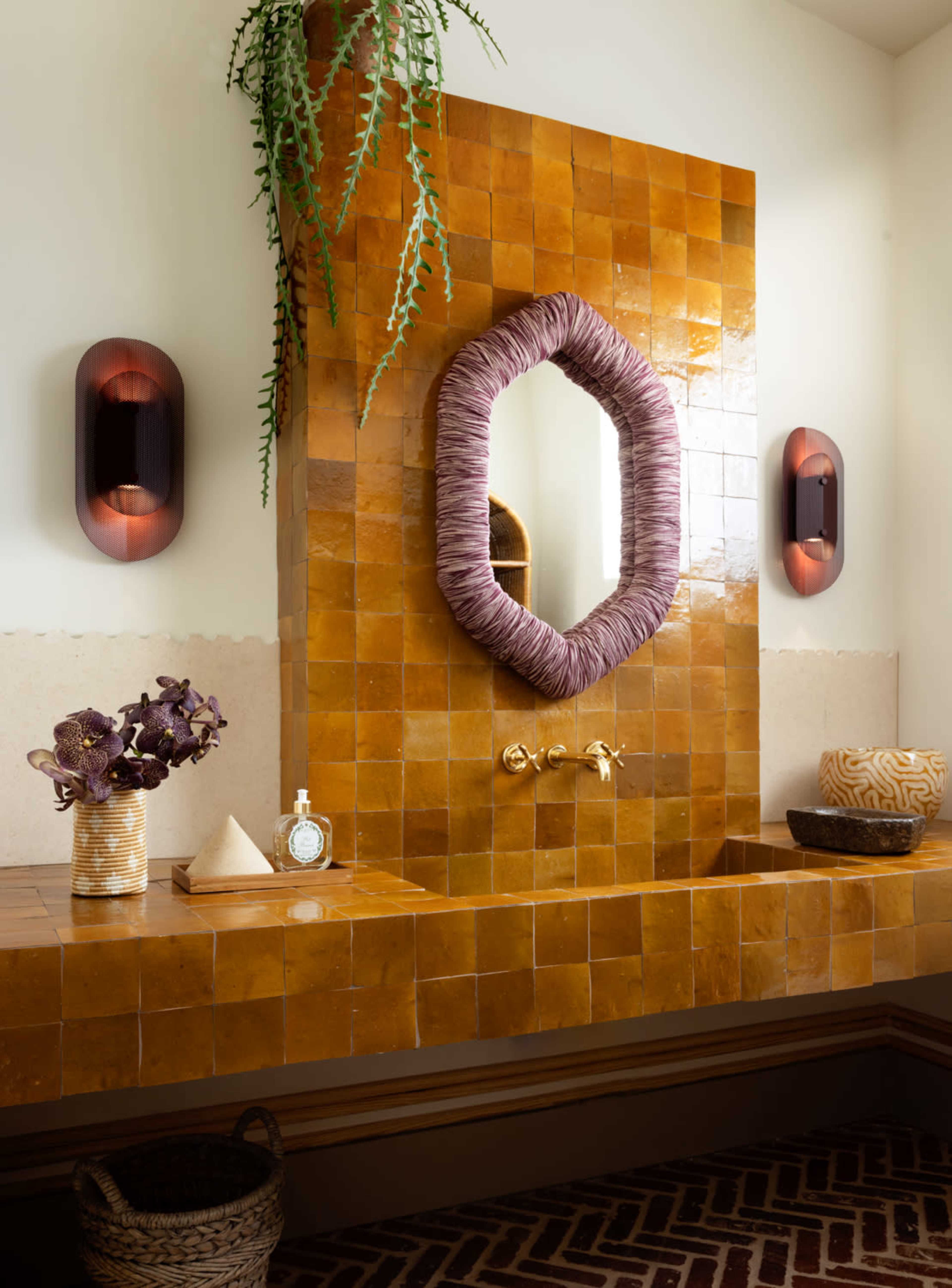 A bathroom features a large, textured tiled wall with an oval mirror, gold fixtures, and decorative plants, accented by modern lighting and vases.