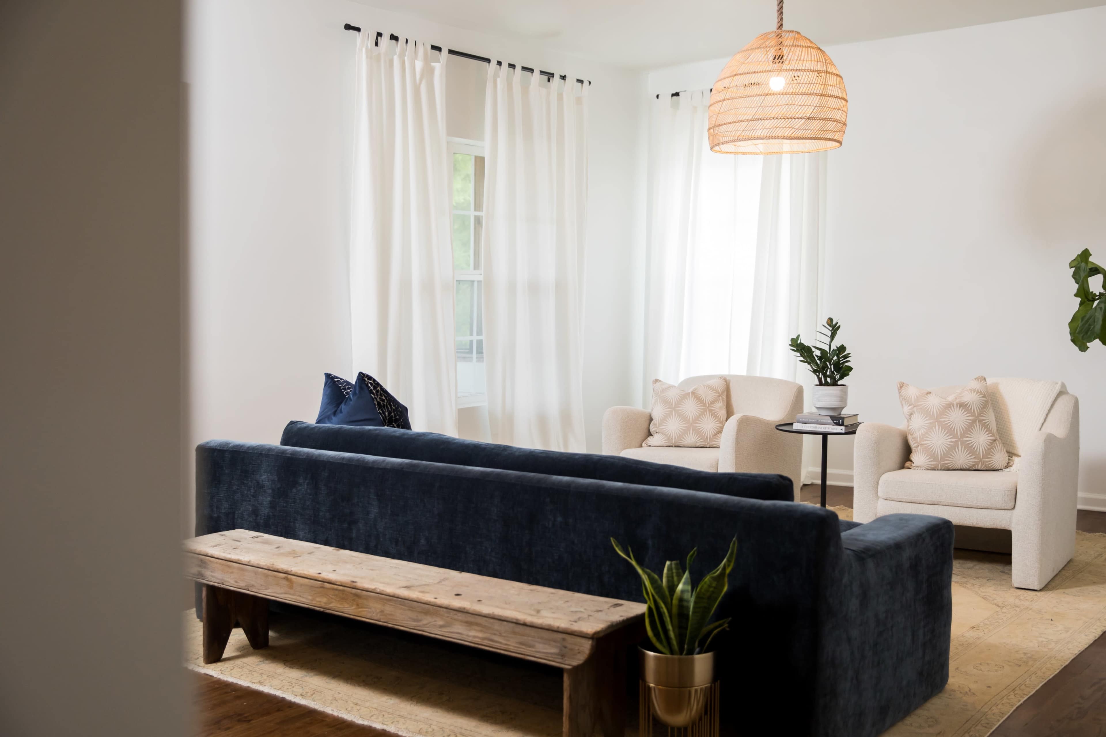 A cozy living room with a dark blue sofa, two white armchairs, a wooden coffee table, and large windows covered with sheer white curtains.
