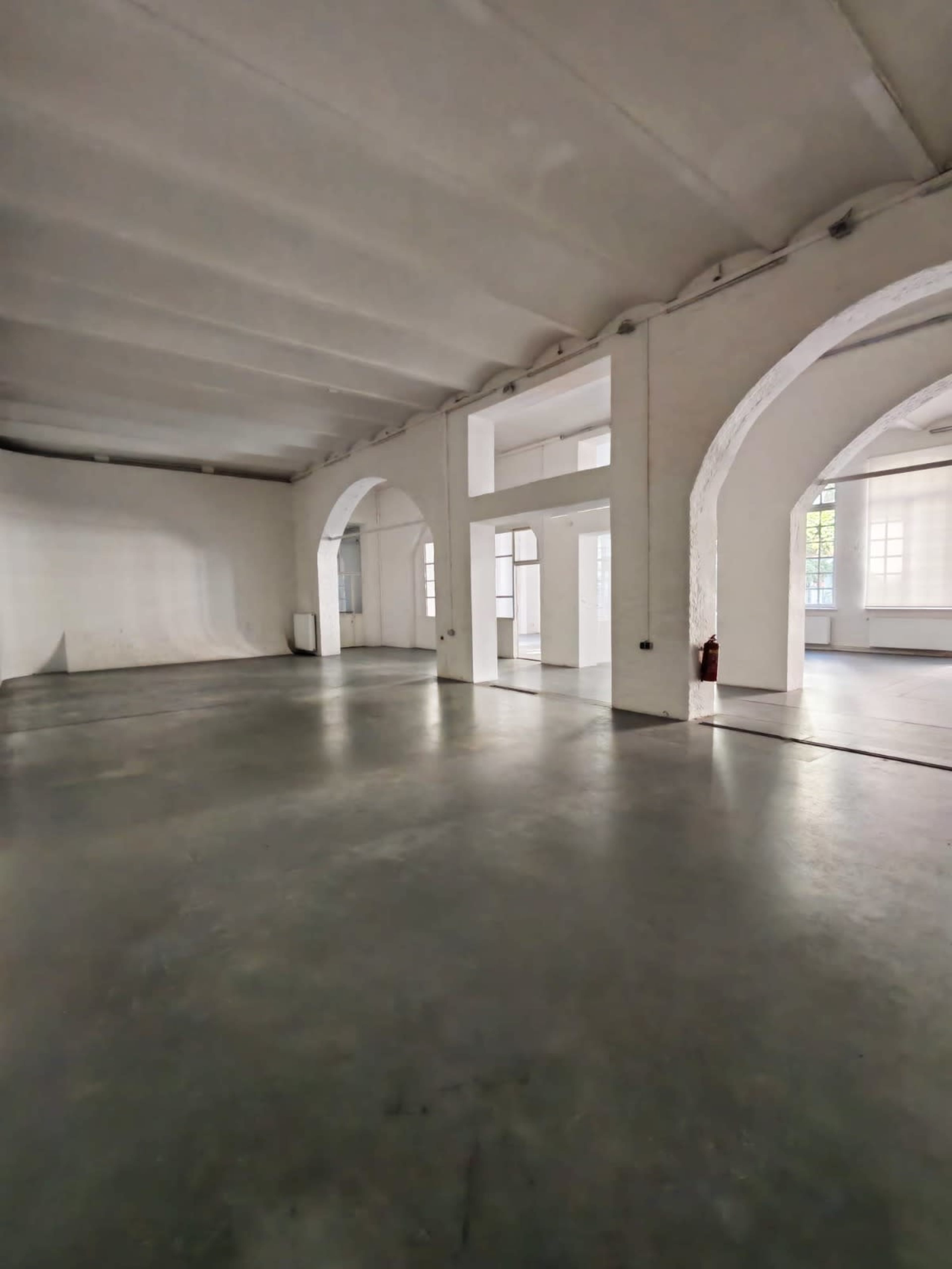 The image shows a spacious, empty room with arched doorways and large windows, featuring a concrete floor and white walls.