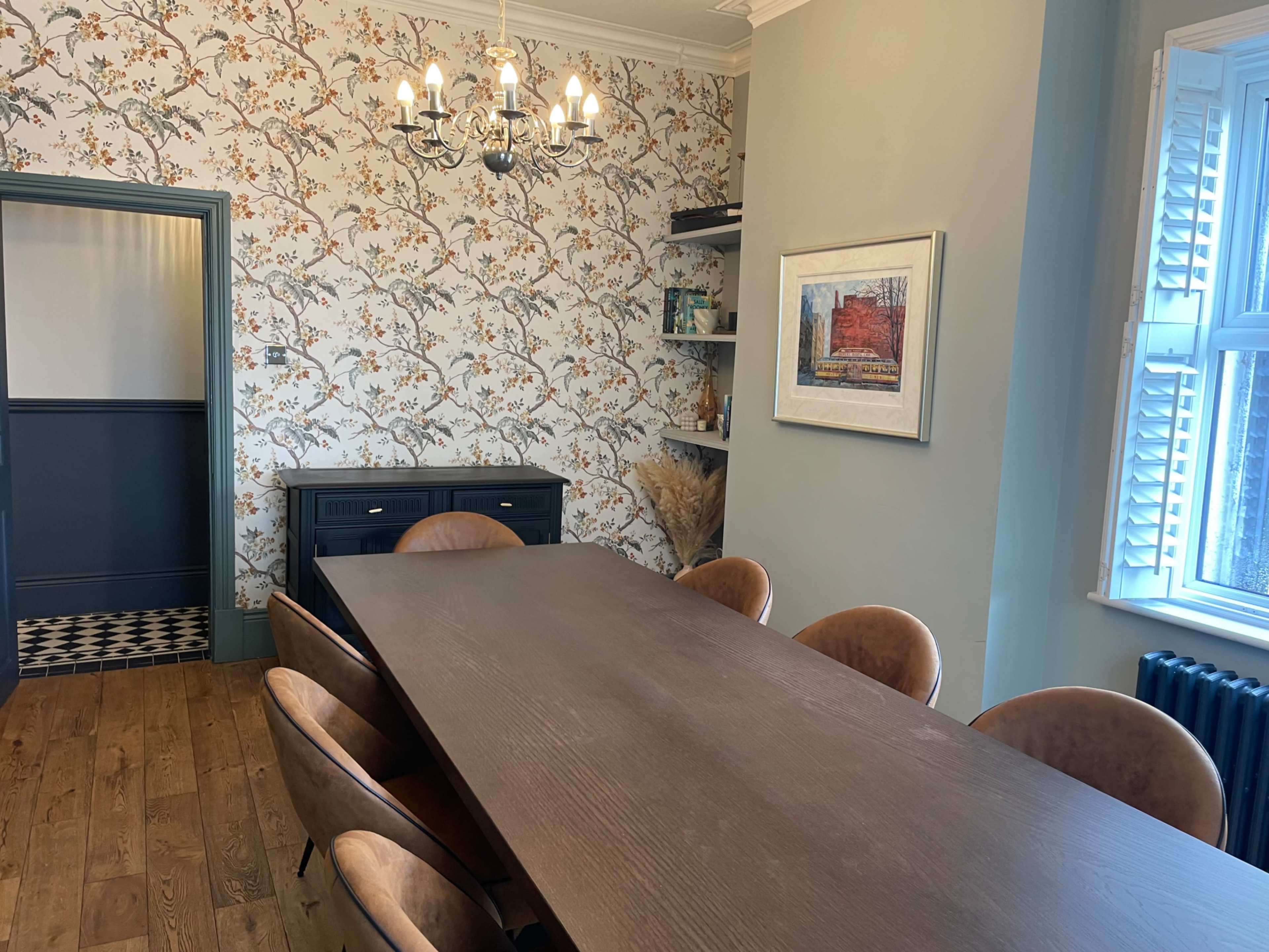 The image shows a dining room with a long wooden table surrounded by round chairs, a floral wallpapered wall, and a chandelier overhead.
