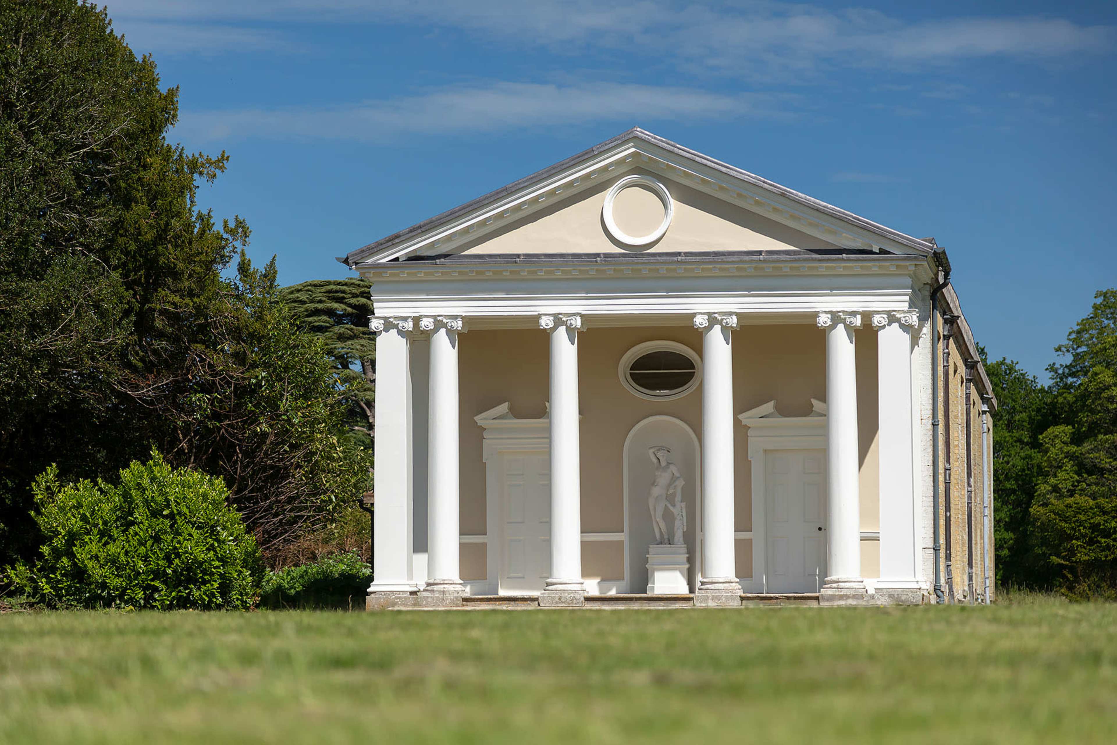 A classical-style building with six columns and a central statue is set against a blue sky and green landscape.