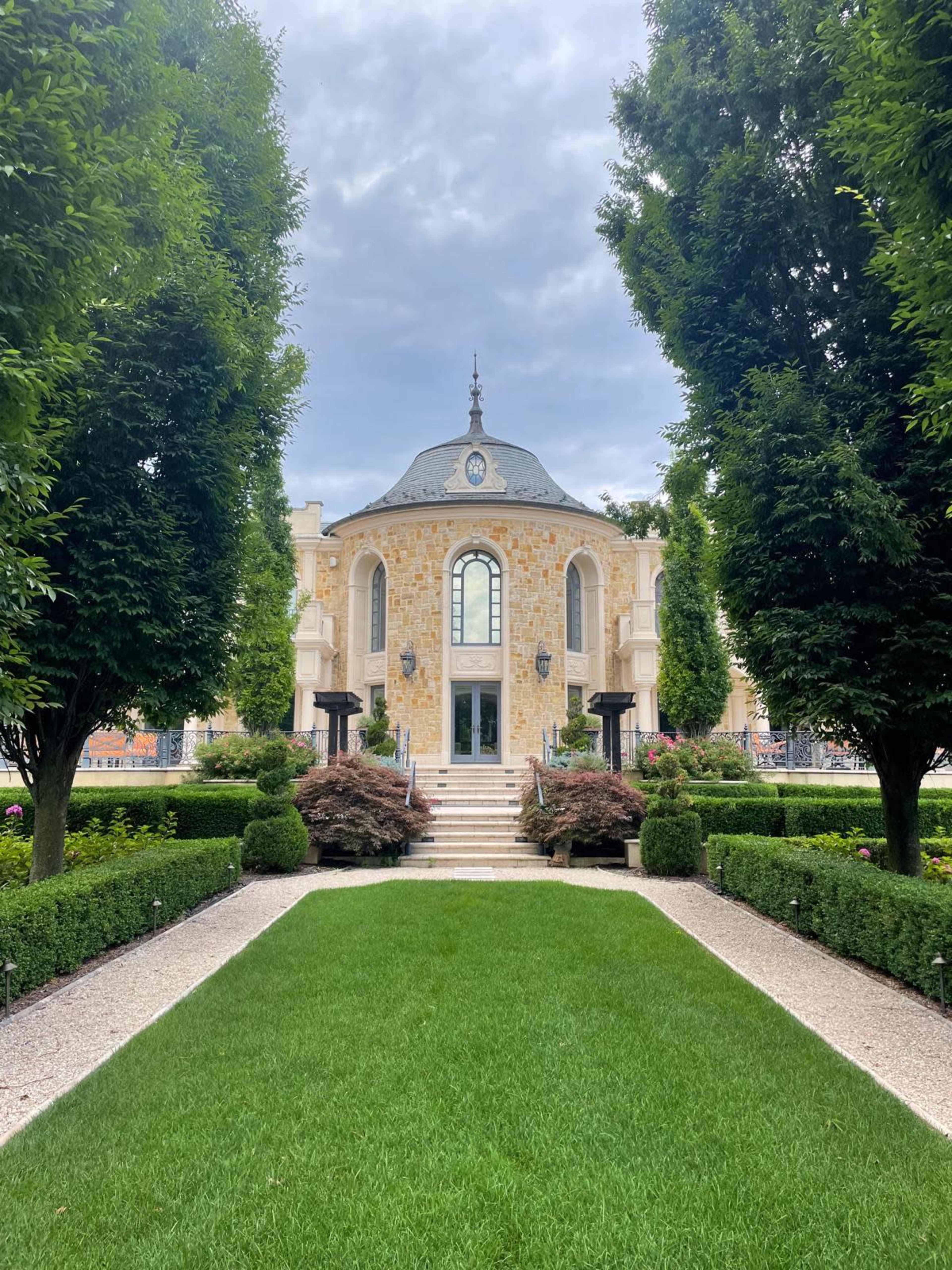 A well-manicured lawn leads to a stone building with a rounded entrance framed by symmetrical hedges and trees.