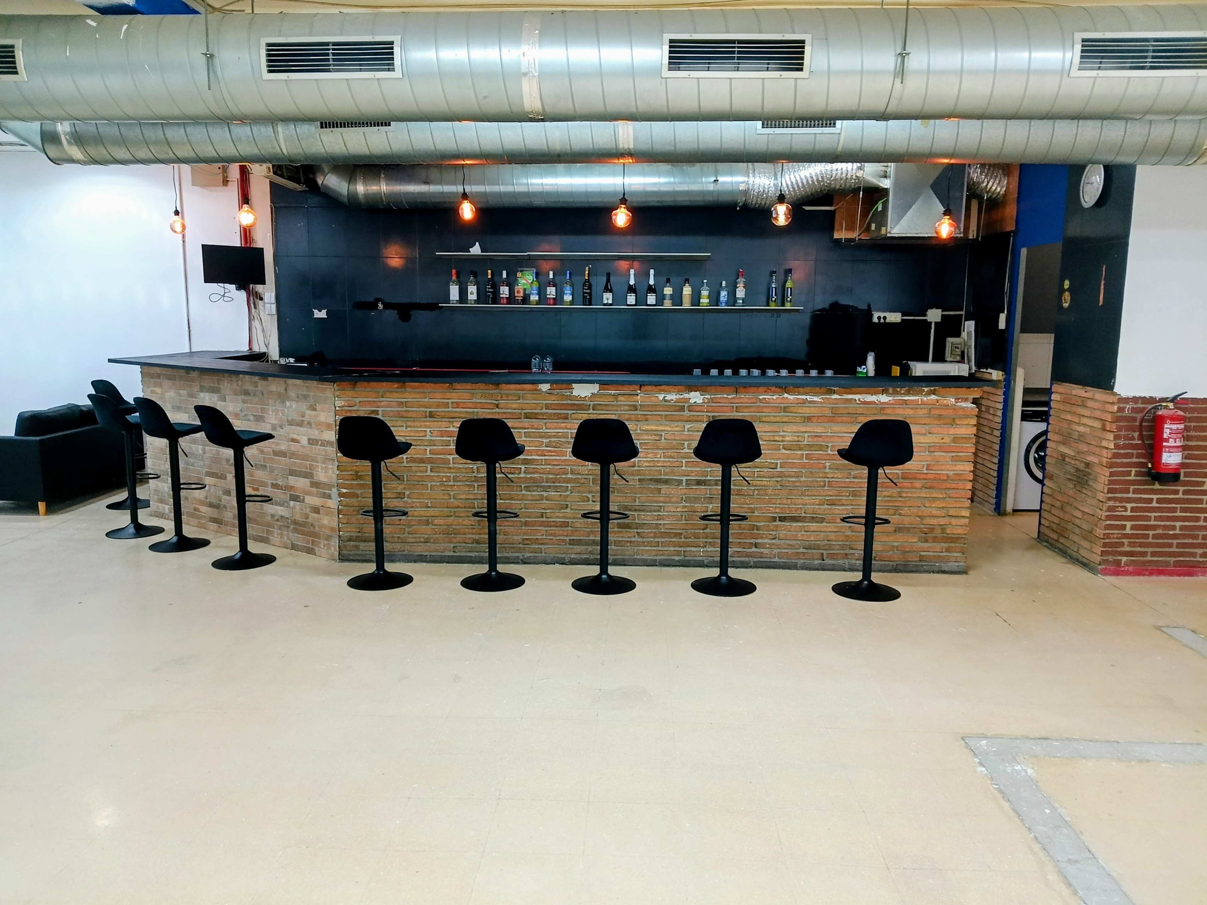 The image shows a modern bar with a stone countertop and several black stools lined up in front, illuminated by pendant lights overhead.