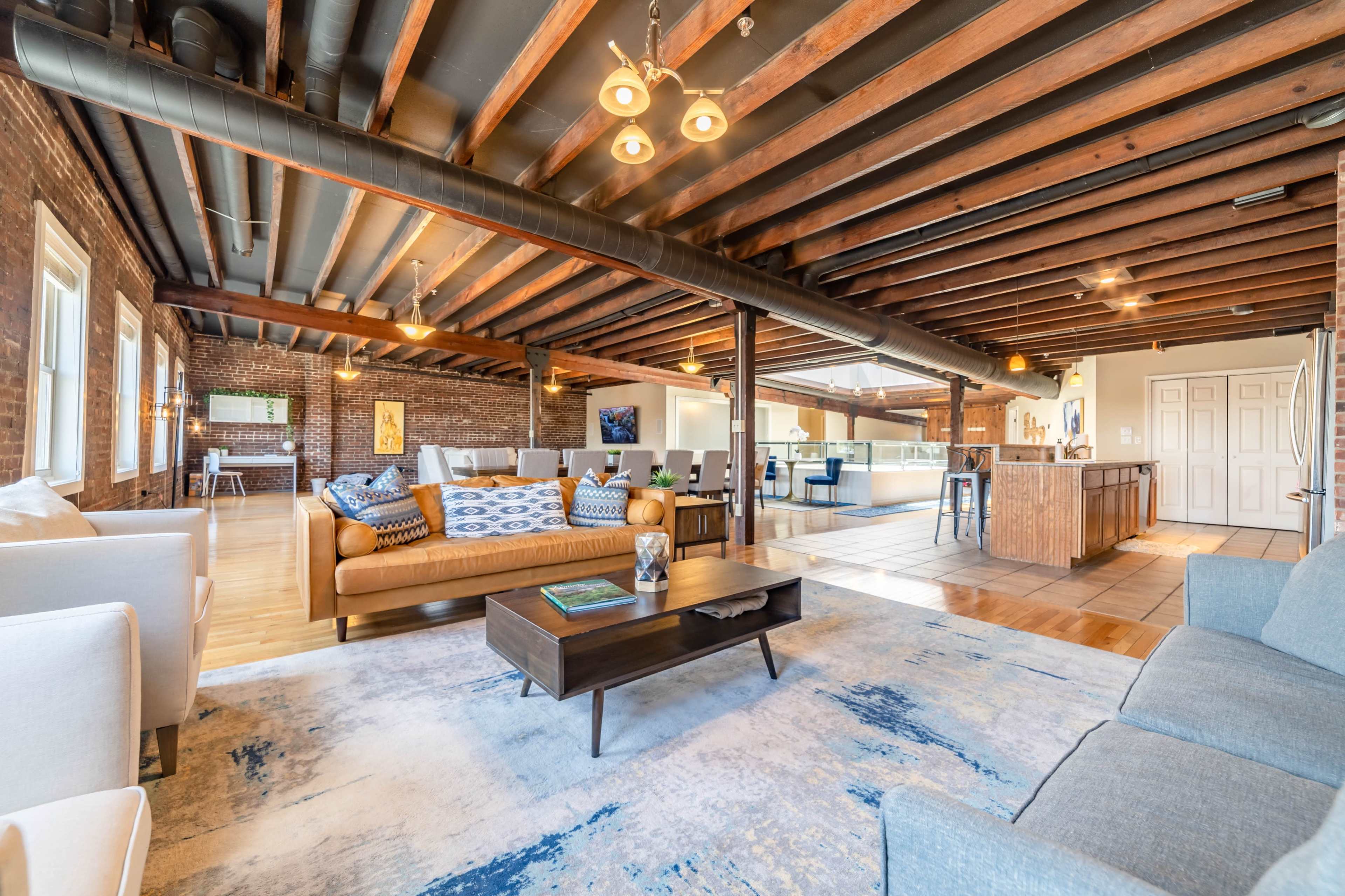 The spacious living area features exposed wooden beams, a mix of comfortable seating arrangements, and a modern kitchen visible in the background.