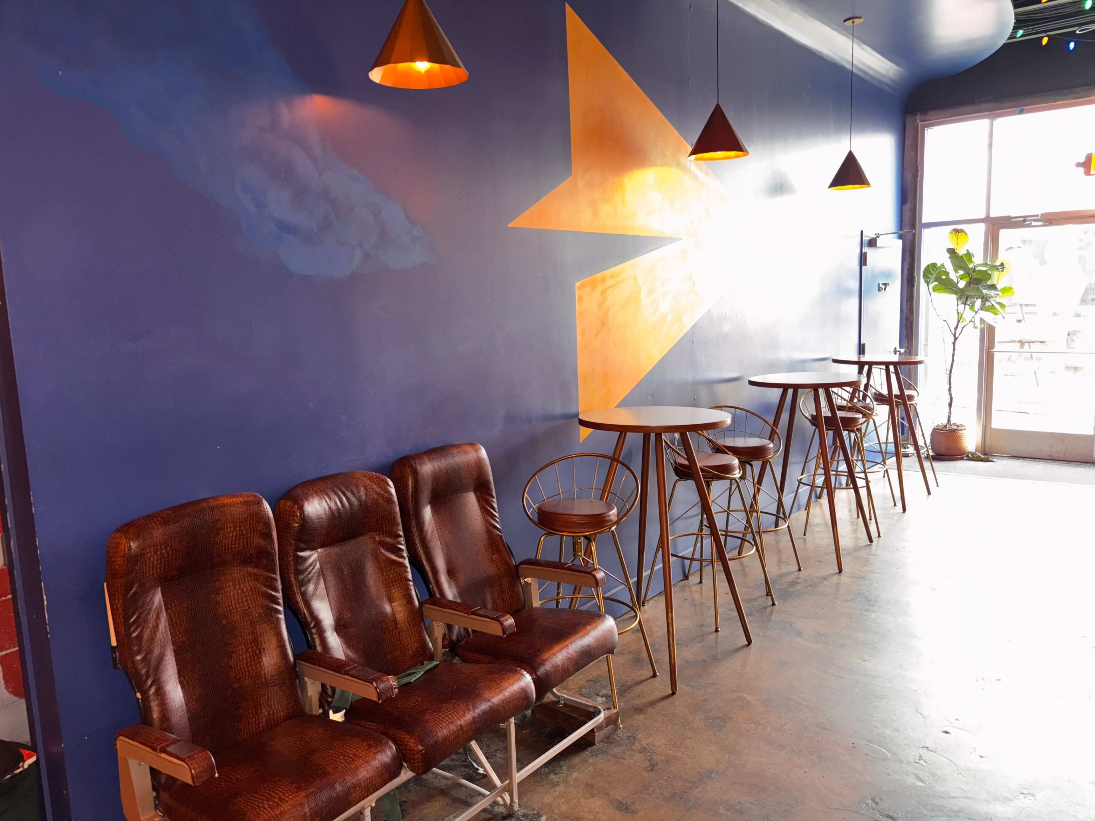 The image shows a cozy interior space with wooden seating, high tables, and a large yellow arrow mural on a blue wall.