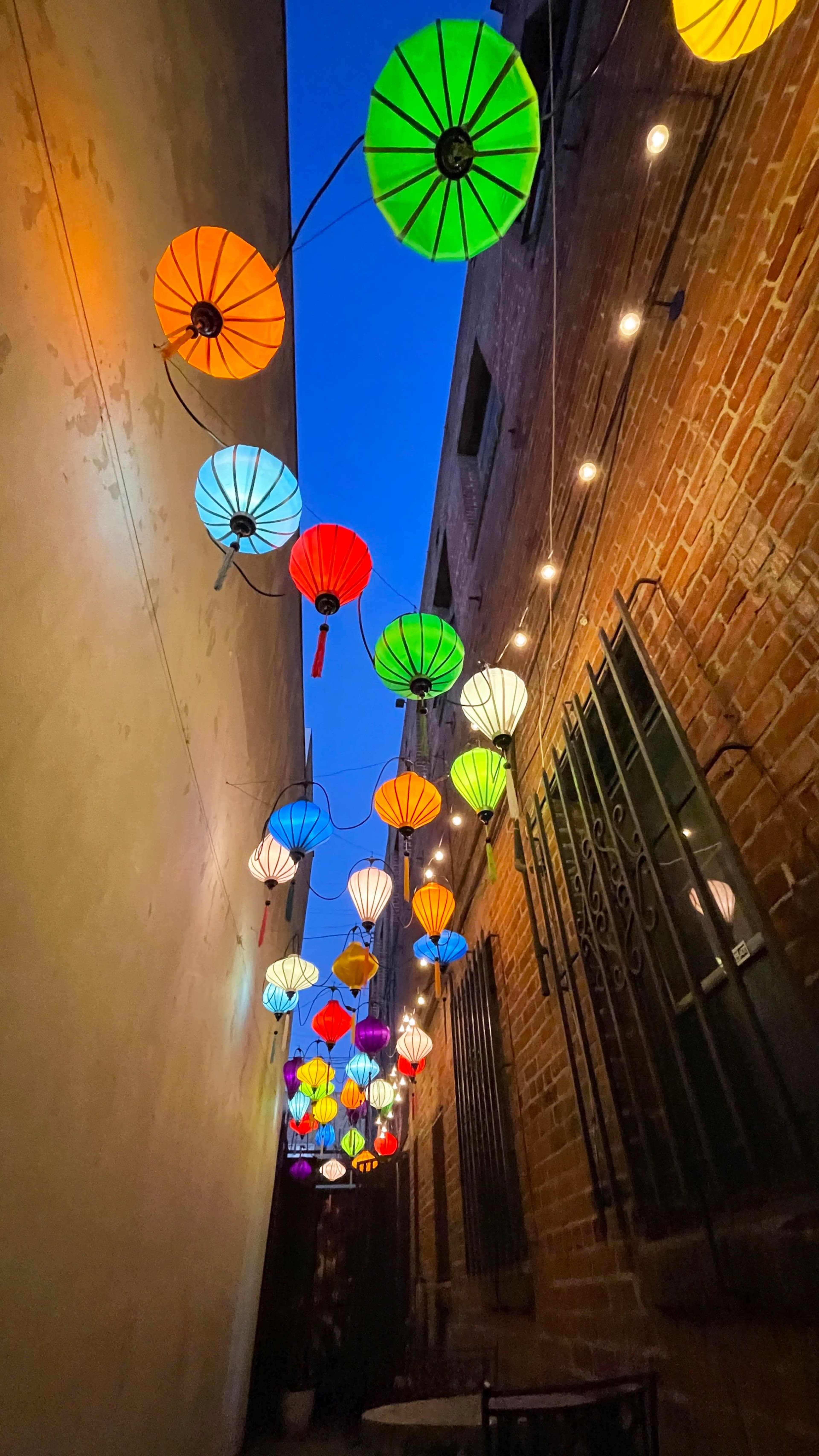 Colorful lanterns hang above an alleyway, casting bright hues against the brick walls and dimming twilight sky.