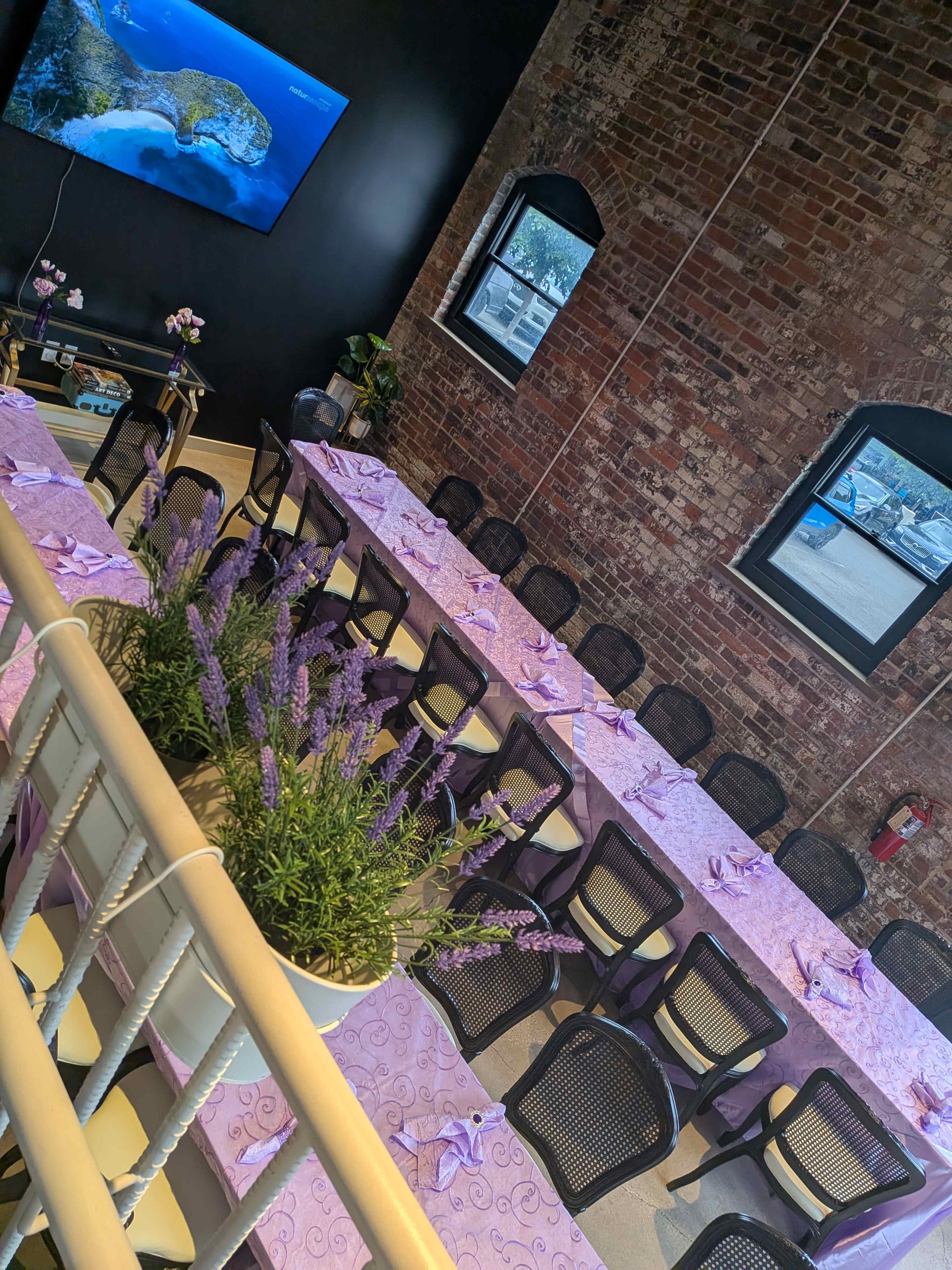 A long dining table with purple tablecloths and napkins is set up in a brick-walled room, while a television displays a scenic view on the wall.