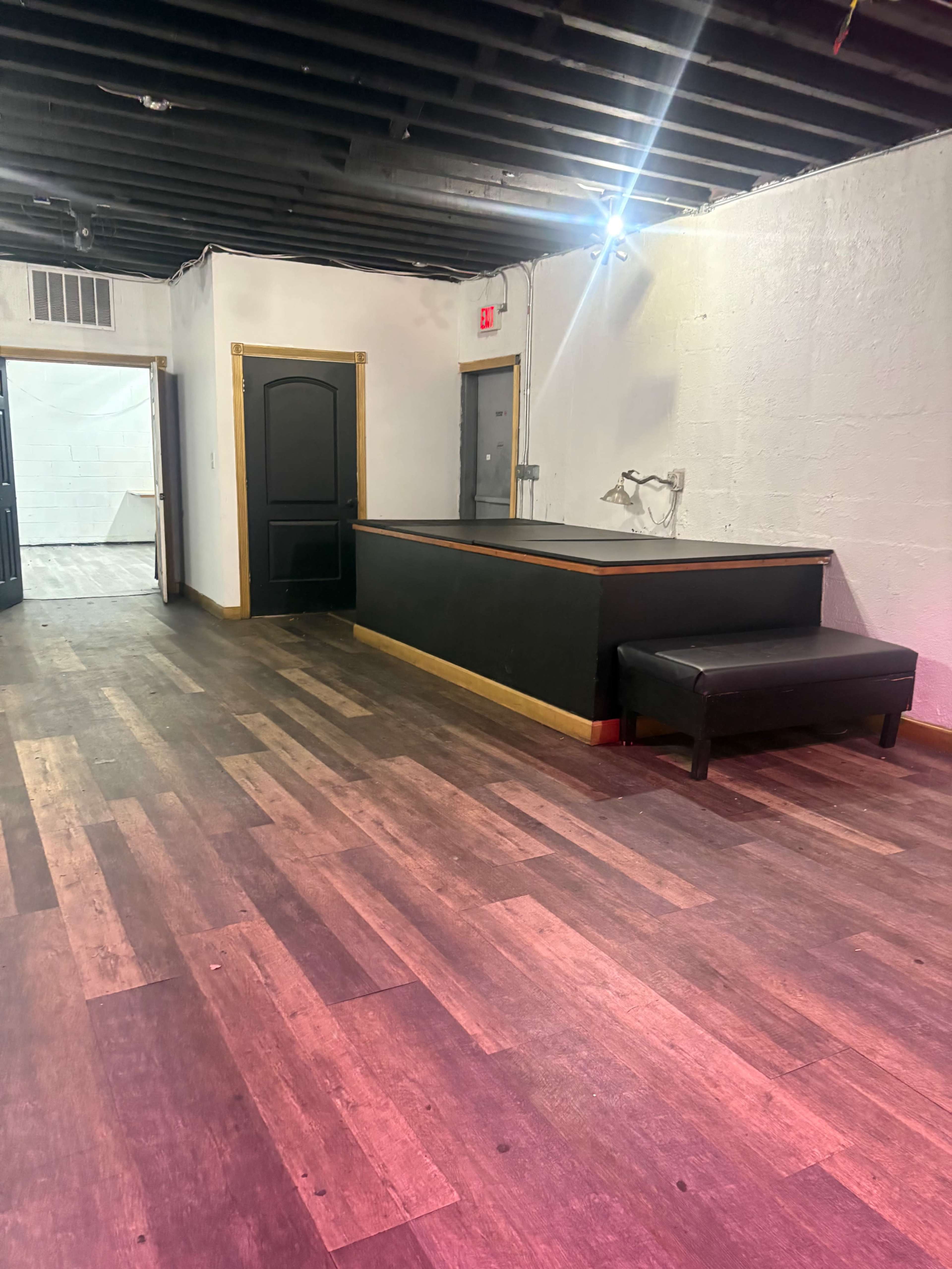 The image shows an empty room with dark wood flooring, a lower black platform against one wall, and two doorways leading to other areas.