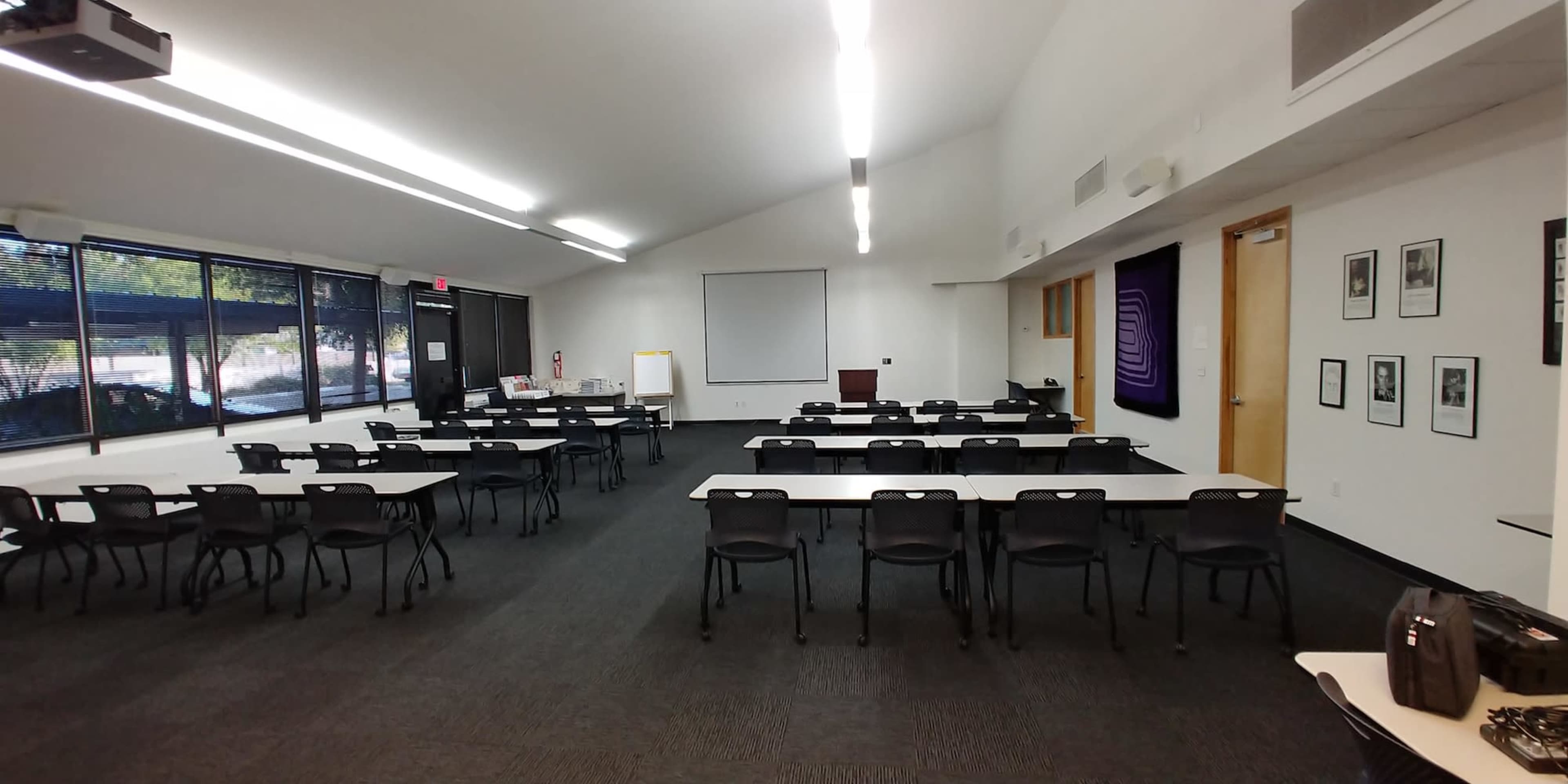 A spacious classroom is arranged with rows of tables and chairs, featuring large windows that allow natural light to enter.