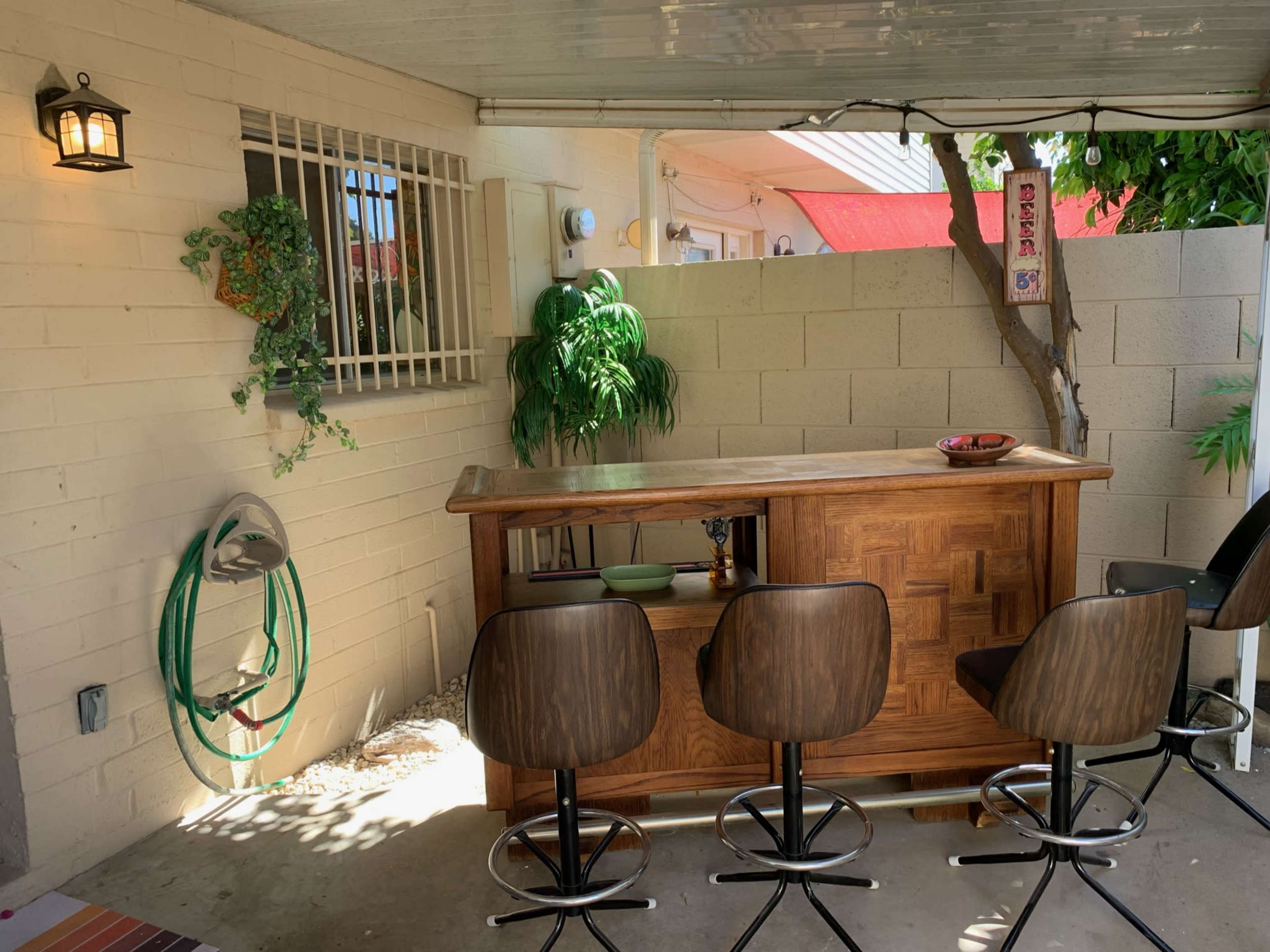 A covered outdoor bar area with three high stools, a wooden bar counter, and a small decorative plant against a block wall.