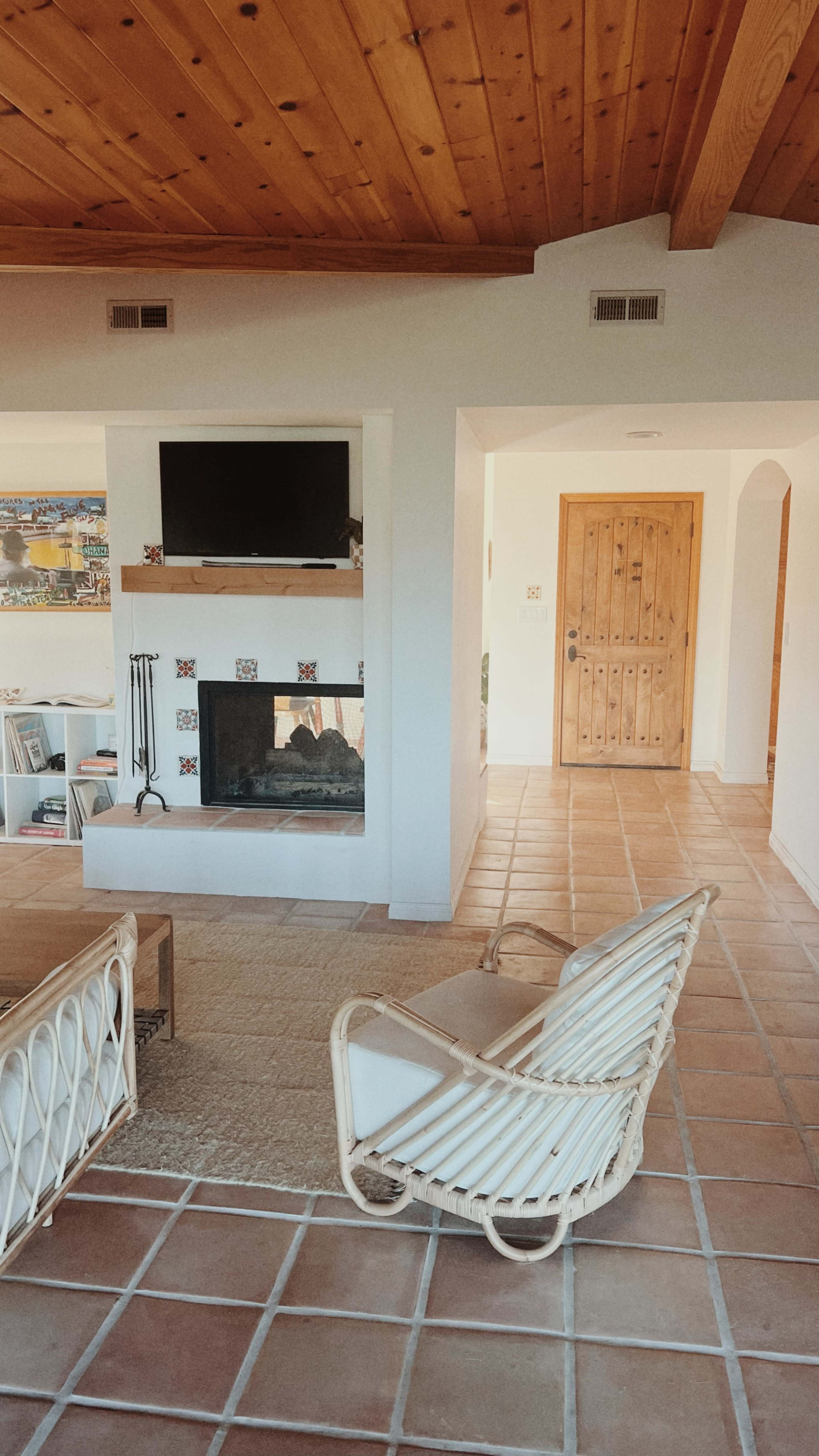 The image shows a living room with a fireplace, a wall-mounted television, a door to the right, and a woven chair placed near a textured area rug on tiled flooring.