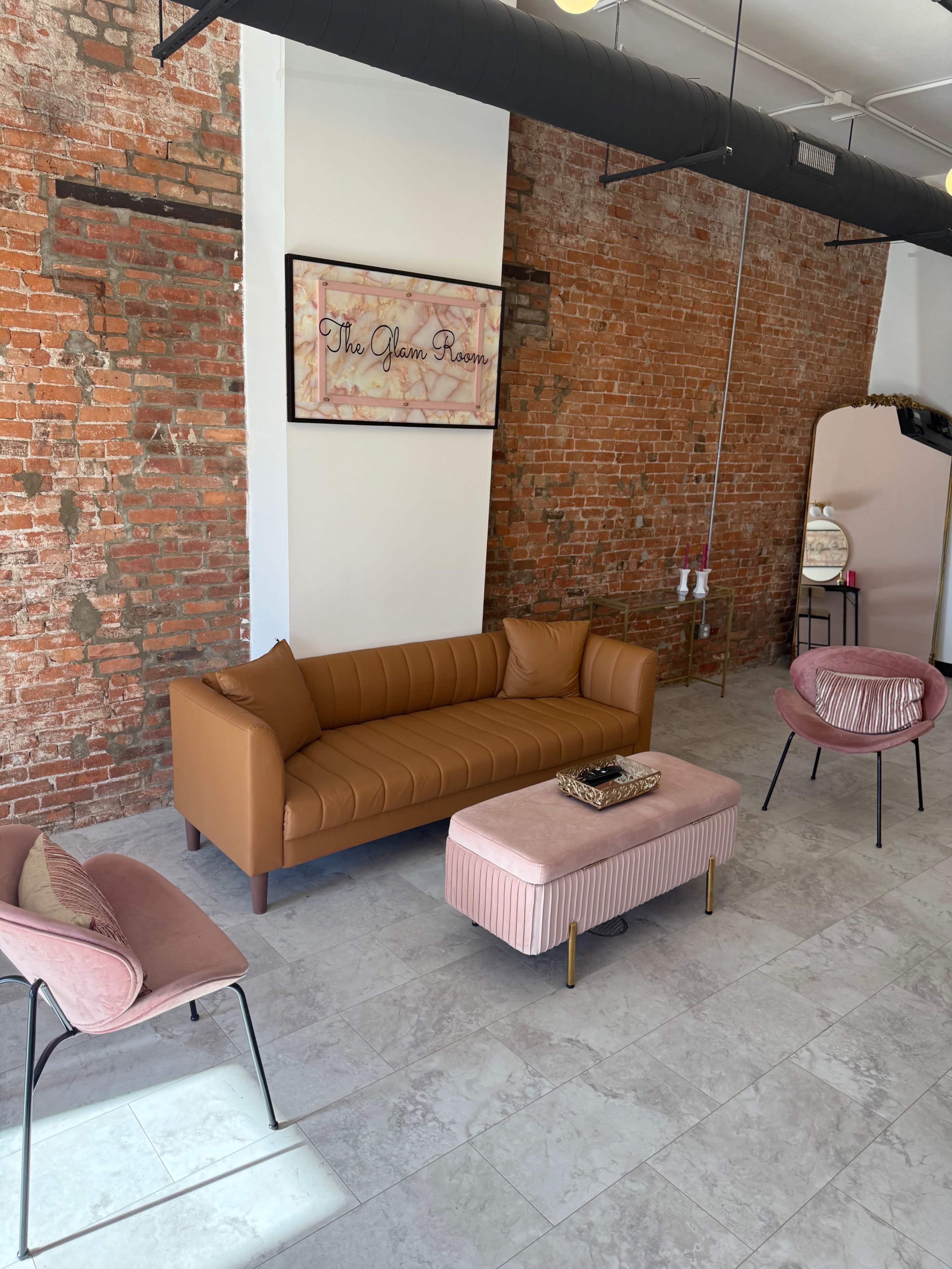 The image shows a stylish seating area featuring a brown couch, a pink ottoman, and two pink chairs against a backdrop of exposed brick walls.