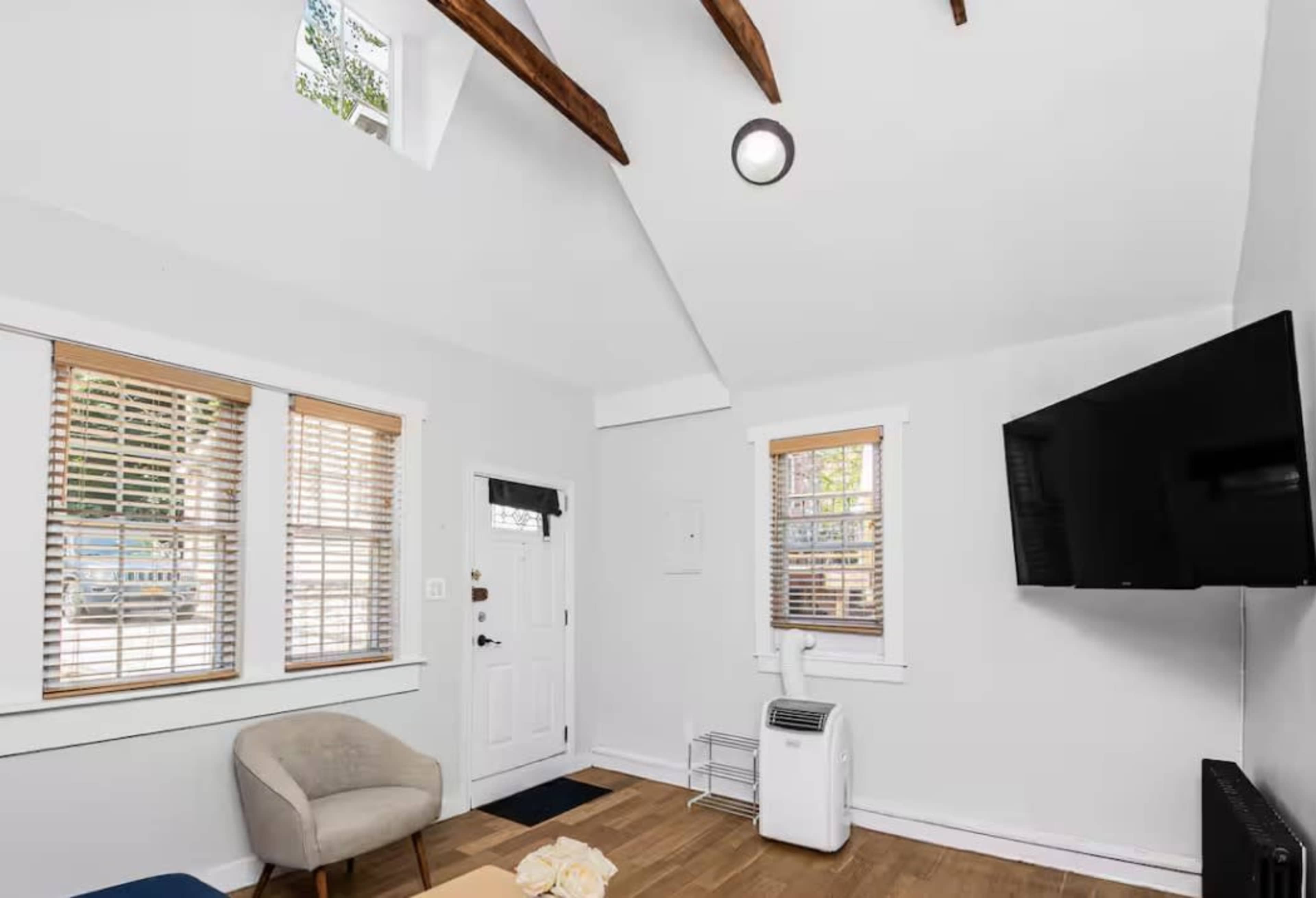 The image shows a bright room with white walls, wooden beams on the ceiling, a small armchair, and a wall-mounted television.
