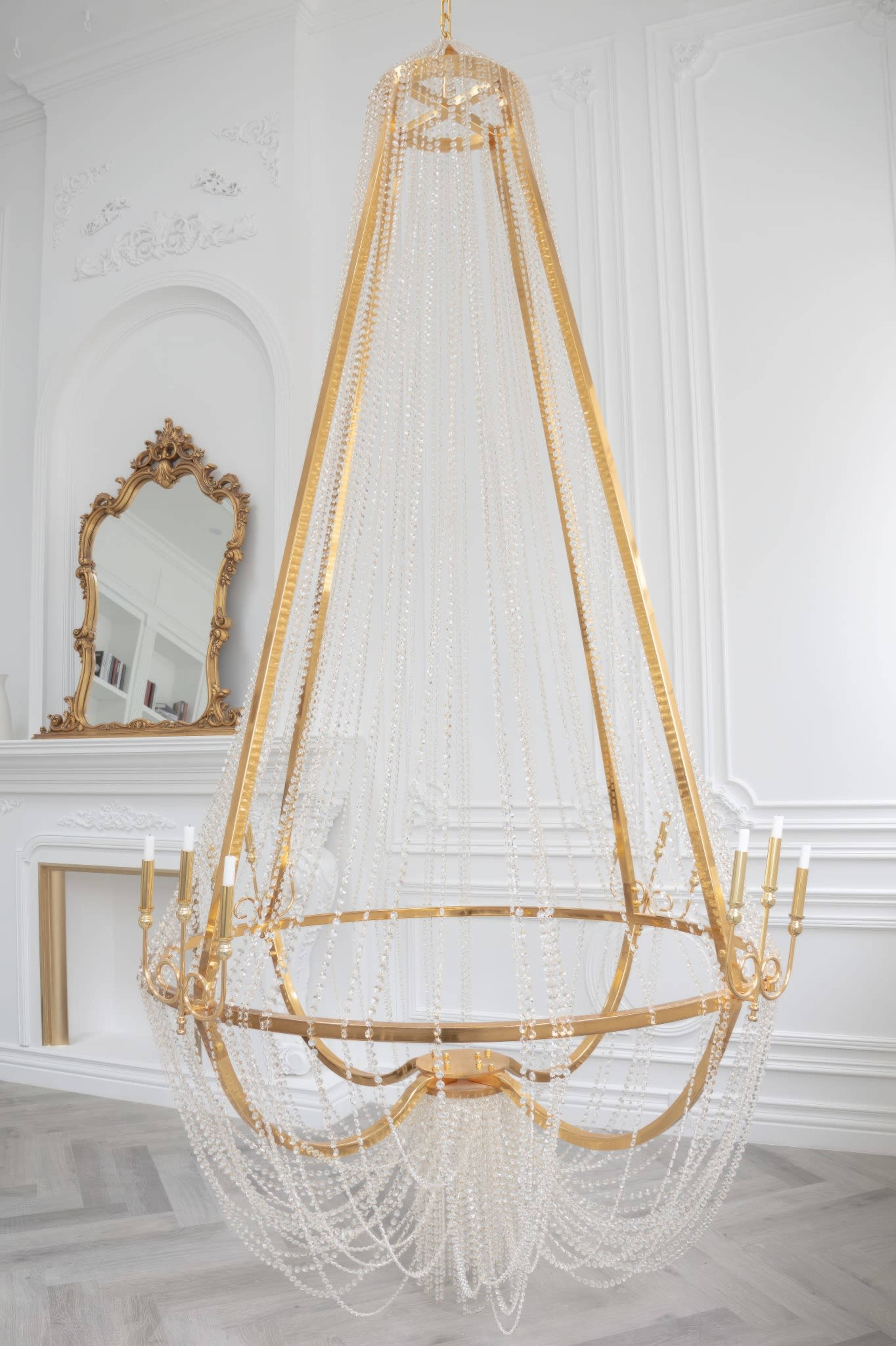 A large, gold and crystal chandelier hangs from the ceiling in a bright, elegantly decorated room with white walls and ornate moldings.