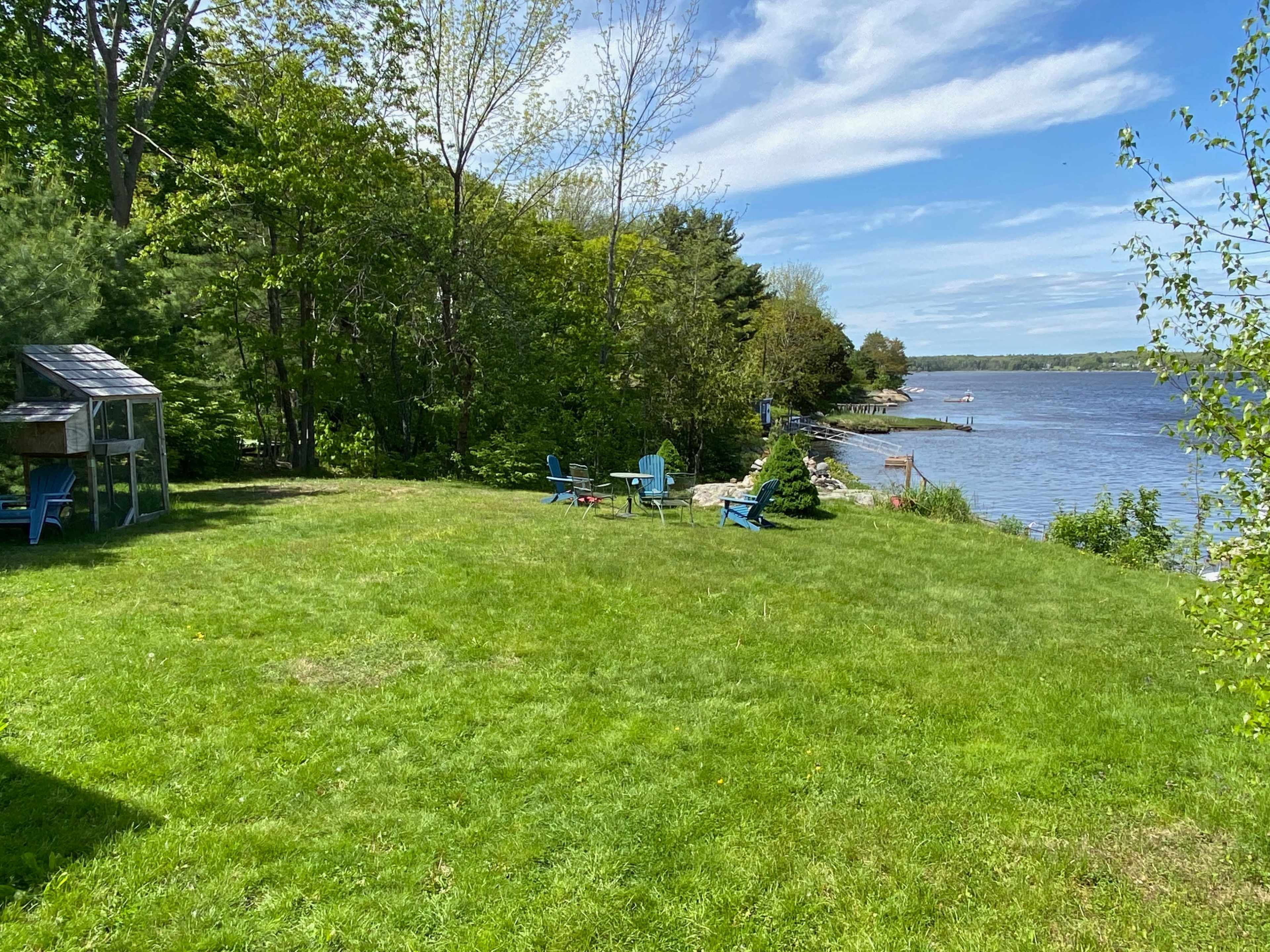 A grassy area near a river features blue Adirondack chairs and a small shed, with trees lining the shore under a clear sky.