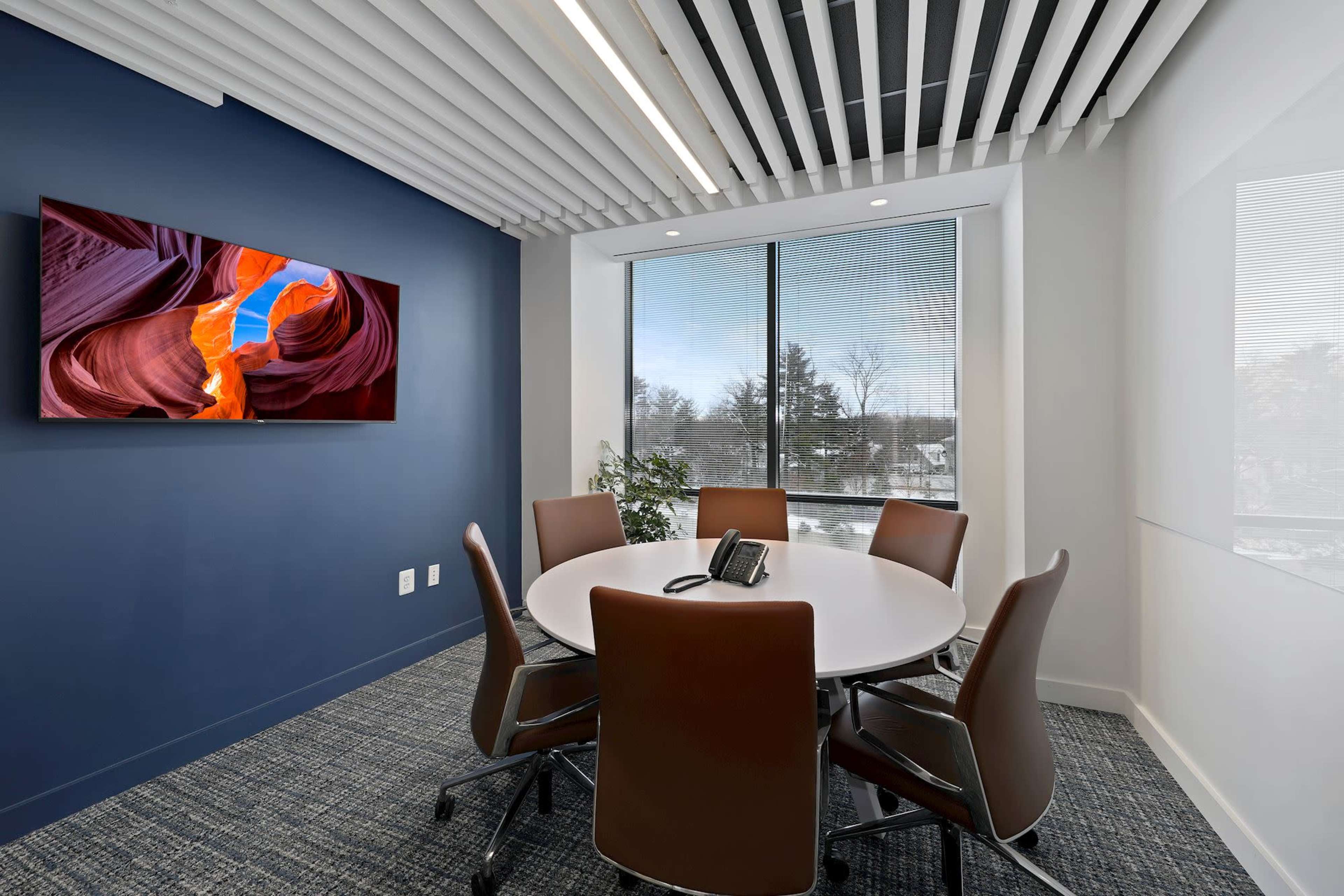 A modern conference room features a round table with six chairs, a wall-mounted TV, and a large window overlooking a exterior landscape.