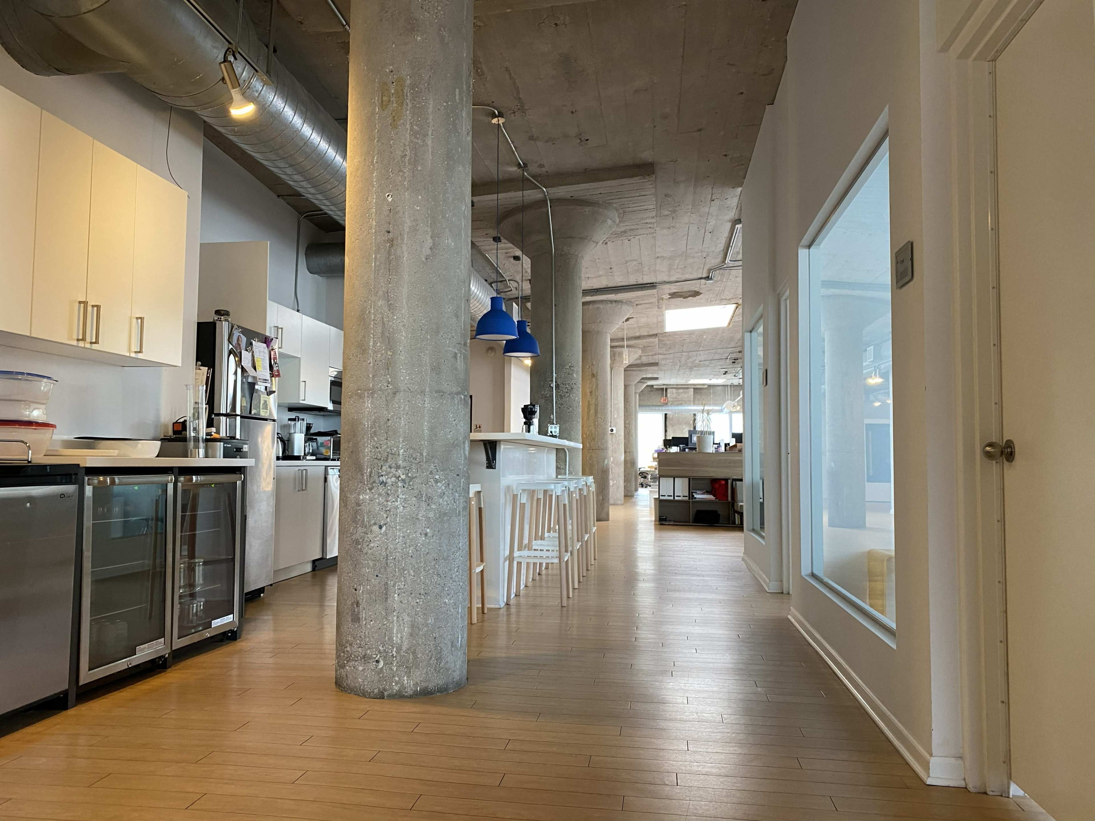 The image shows a spacious, modern kitchen area with concrete pillars and white cabinetry, leading to a bright hallway with additional kitchen space at the end.