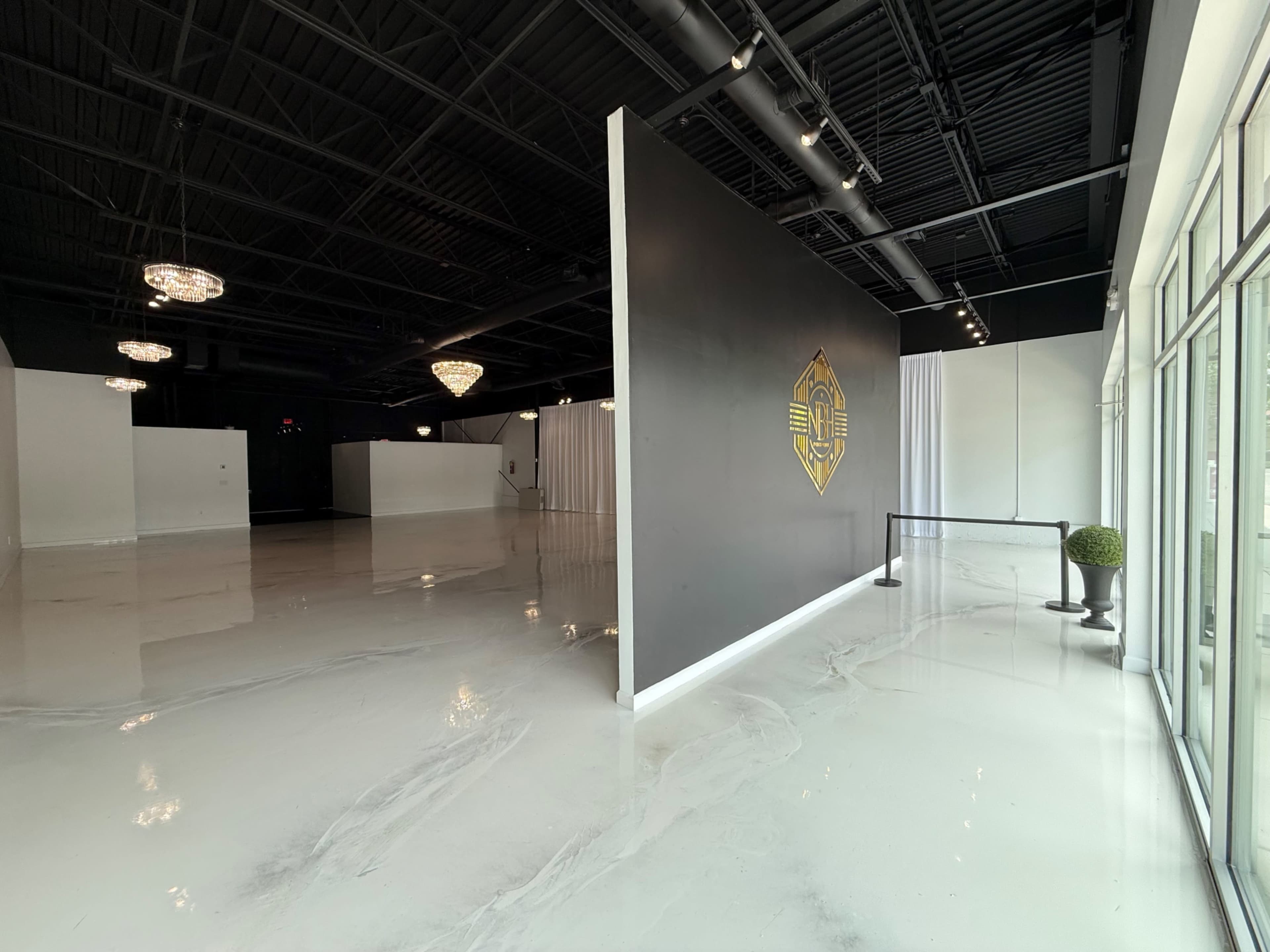 The image shows a spacious, empty indoor venue with a polished white floor, a large black wall featuring a design, and chandeliers hanging from the ceiling.