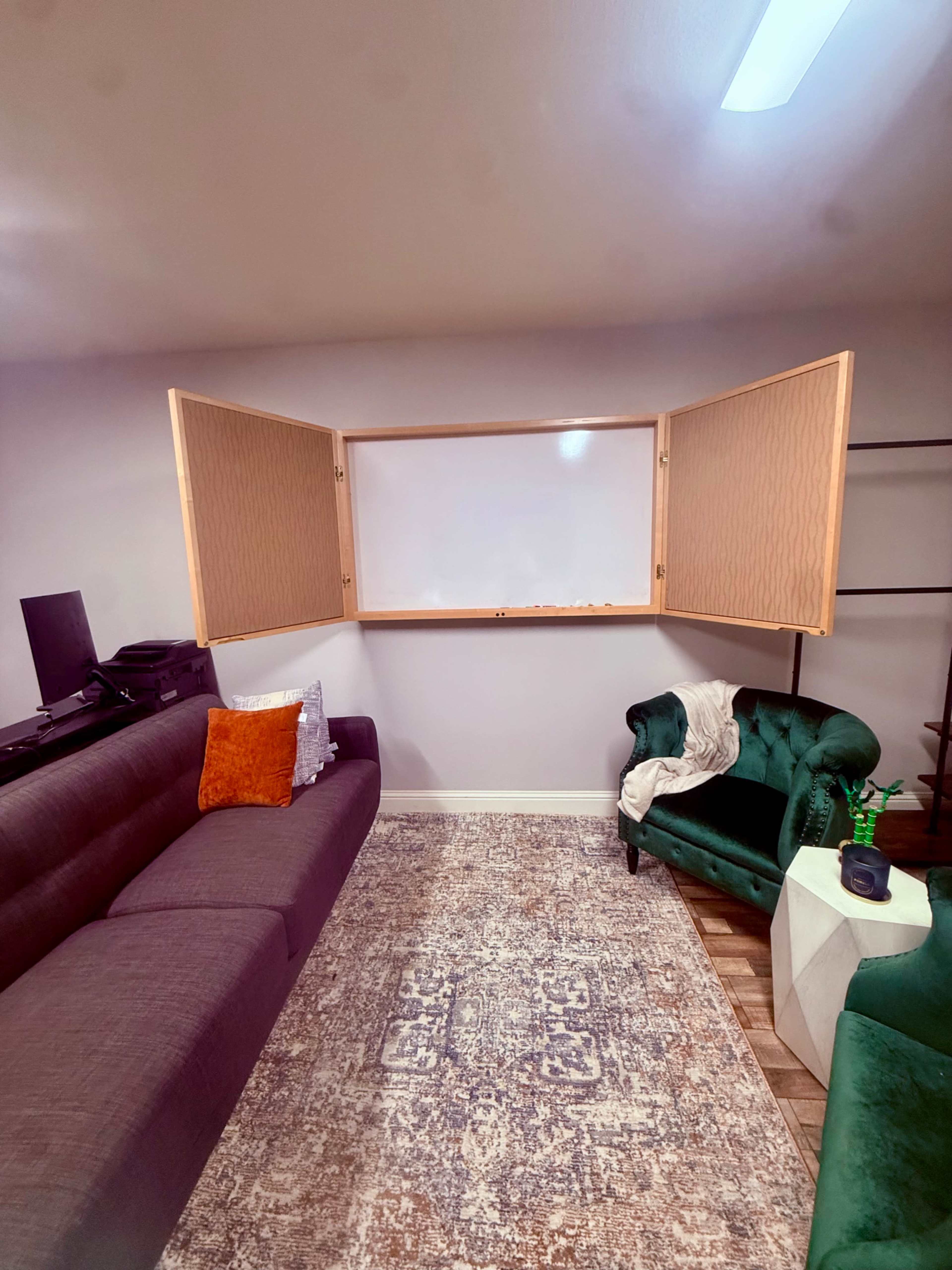 The image shows a cozy room featuring a dark gray sectional sofa, a green upholstered armchair, and a whiteboard mounted in a wooden frame on the wall opposite the seating area.