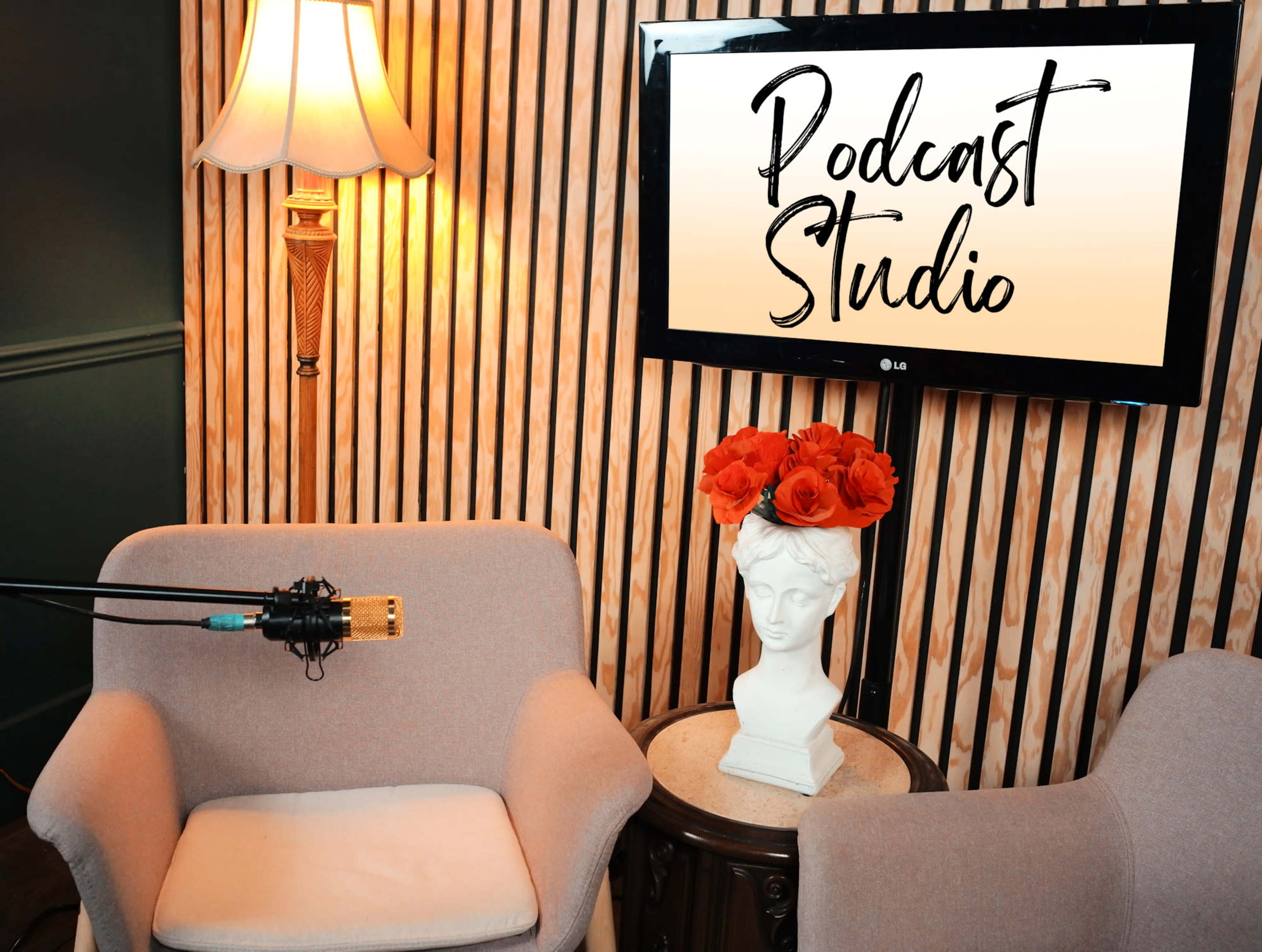 The image shows a cozy podcast studio with two chairs, a microphone setup, and a wall-mounted television displaying "Podcast Studio" alongside a decorative vase of flowers.
