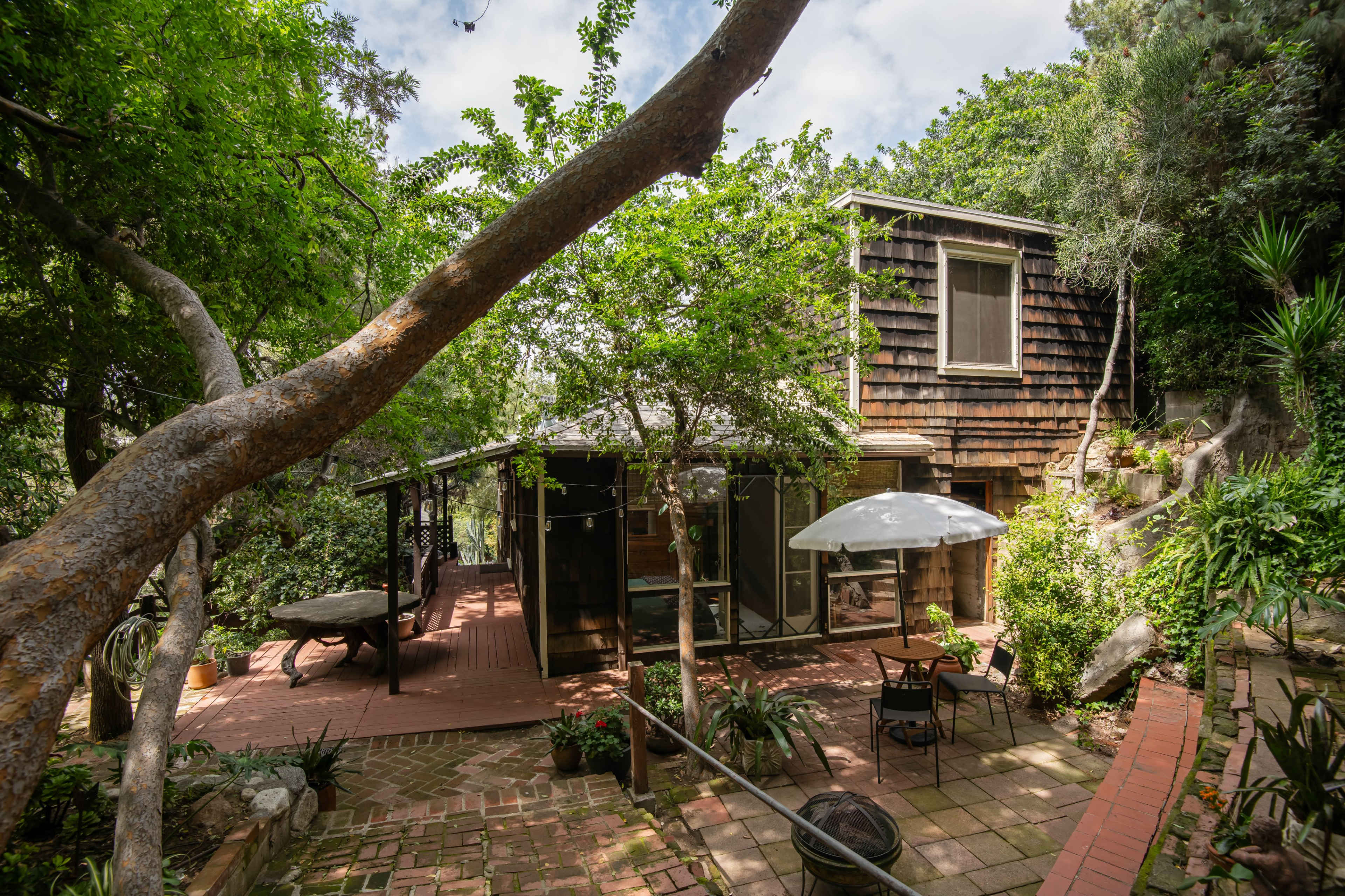 A rustic wooden house is surrounded by greenery and features a patio with an umbrella and seating area.