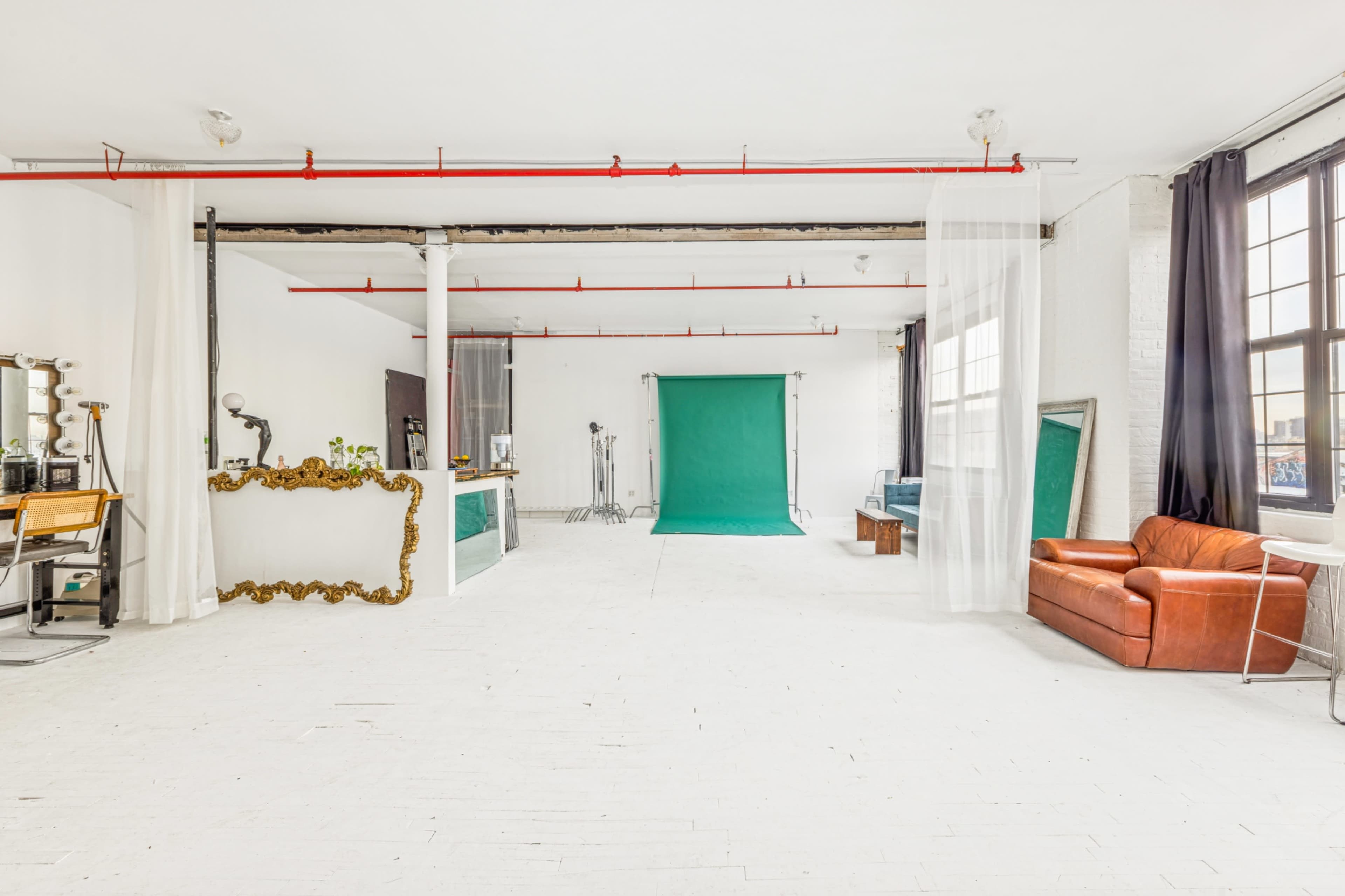The image shows a spacious, bright studio with white walls, hardwood floors, a green backdrop, and various furniture pieces including a brown leather couch and a vintage mirror.
