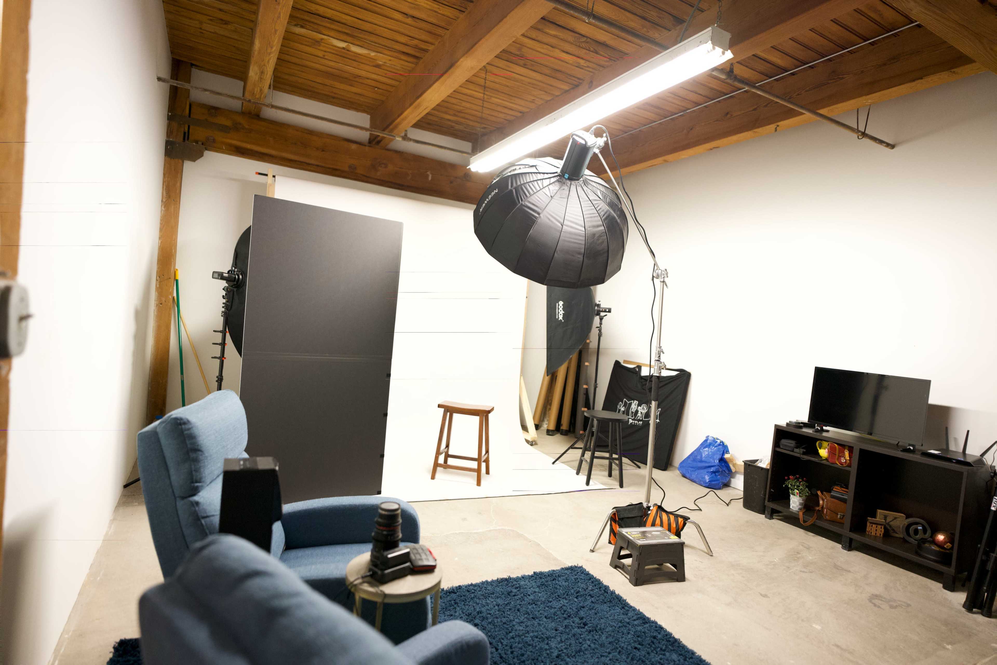 A photography studio with two blue chairs, a table, a large light source, a backdrop, and various props arranged along the walls.