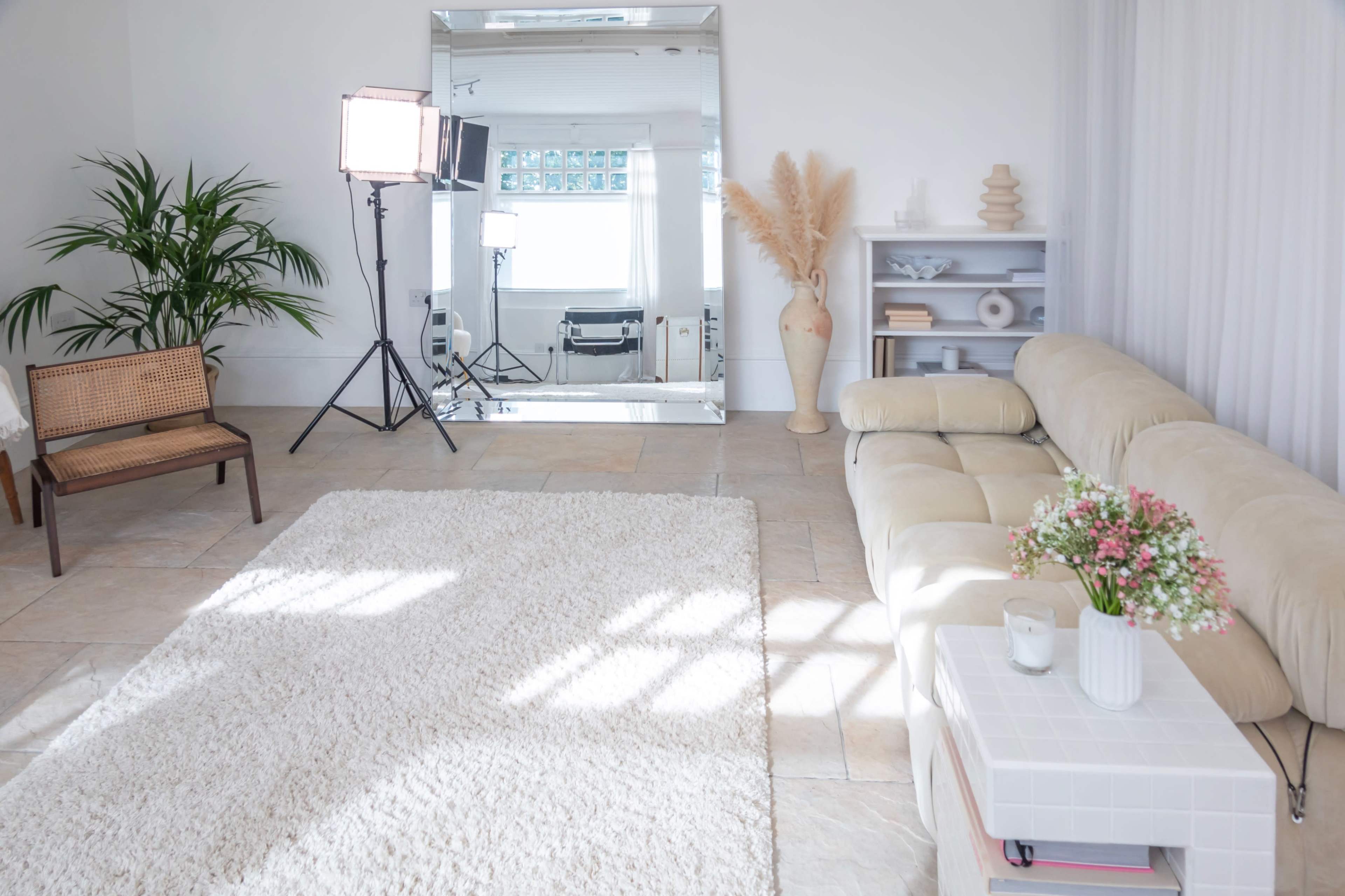 A bright living space features a light-colored sofa, a large mirror, scattered plants, and soft furnishings on a tiled floor.