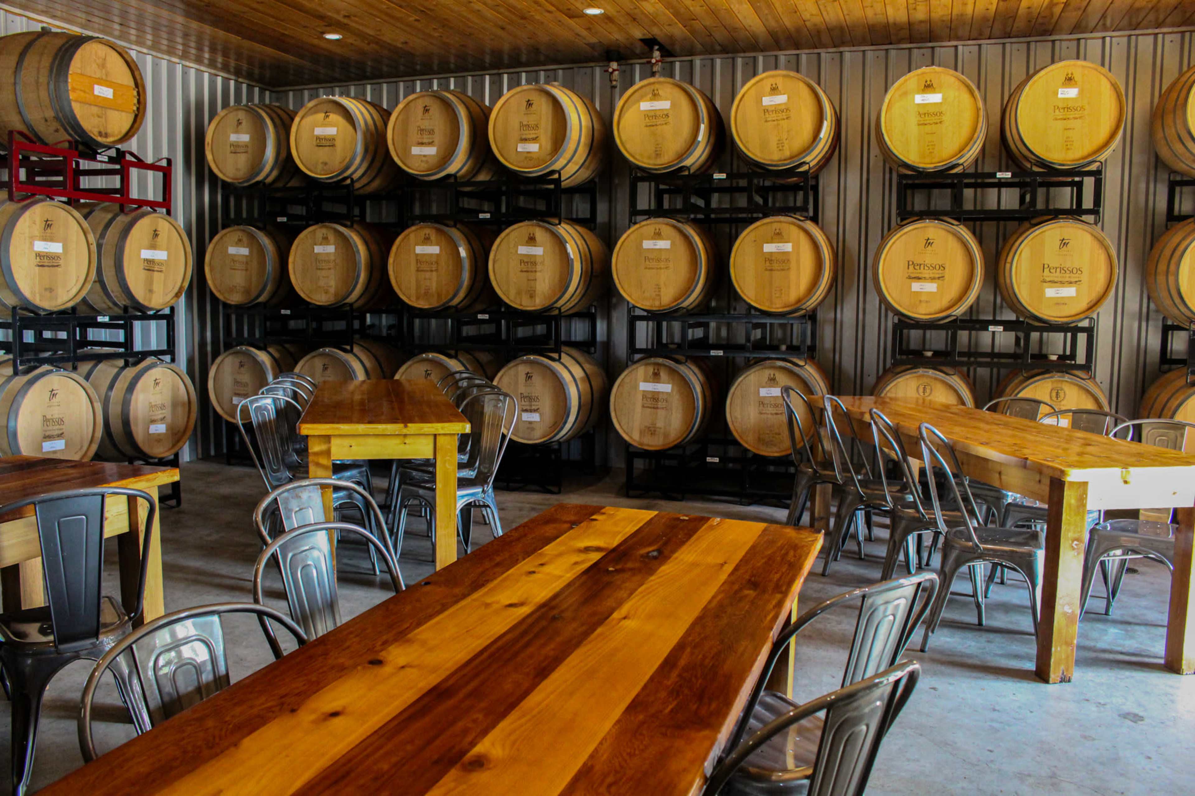 A rustic tasting room with wooden tables and metal chairs, lined with barrels stored on shelves in the background.