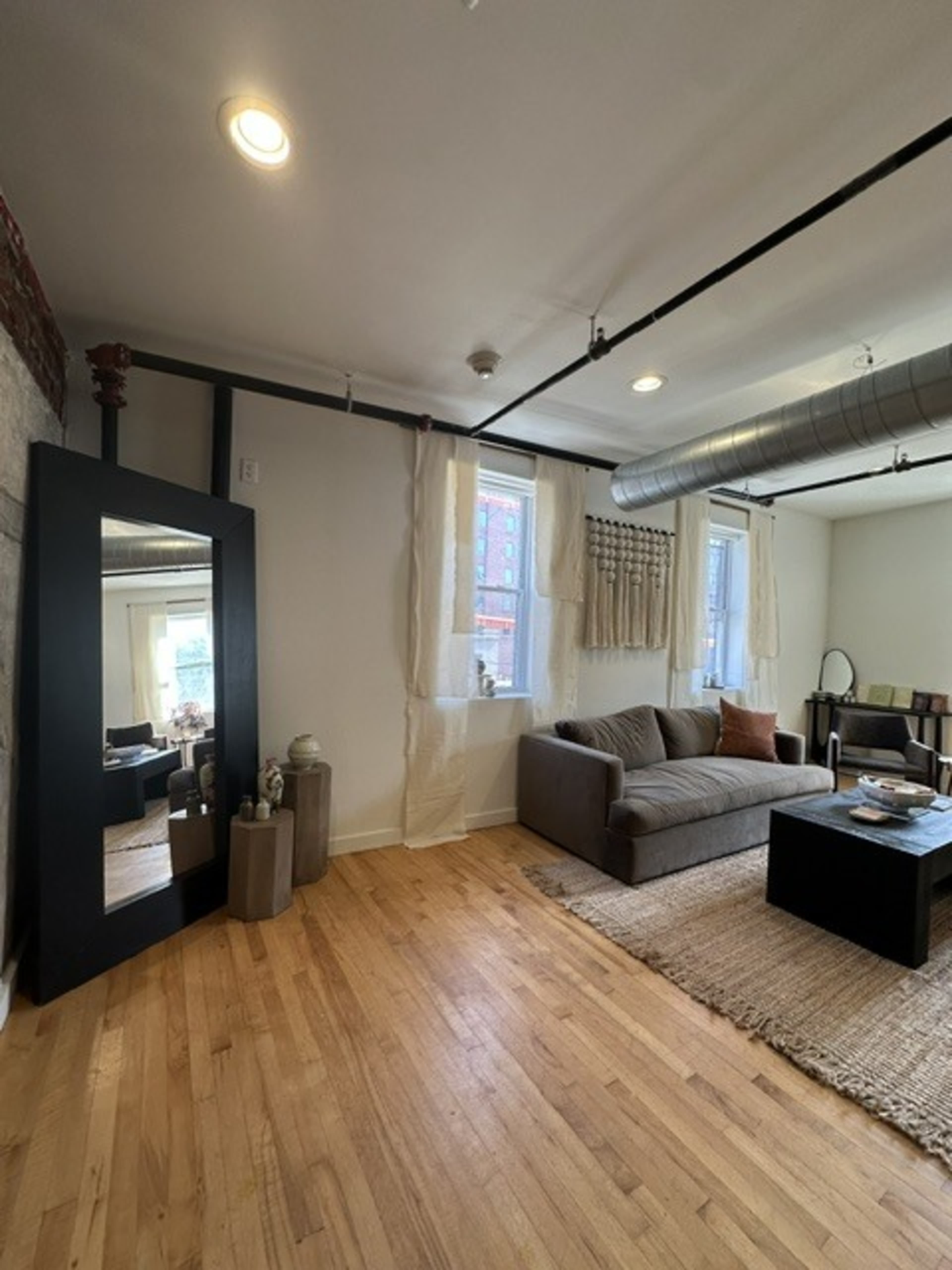 The image shows a bright living room featuring a gray sofa, a coffee table with decor, a large mirror, and windows with sheer curtains.