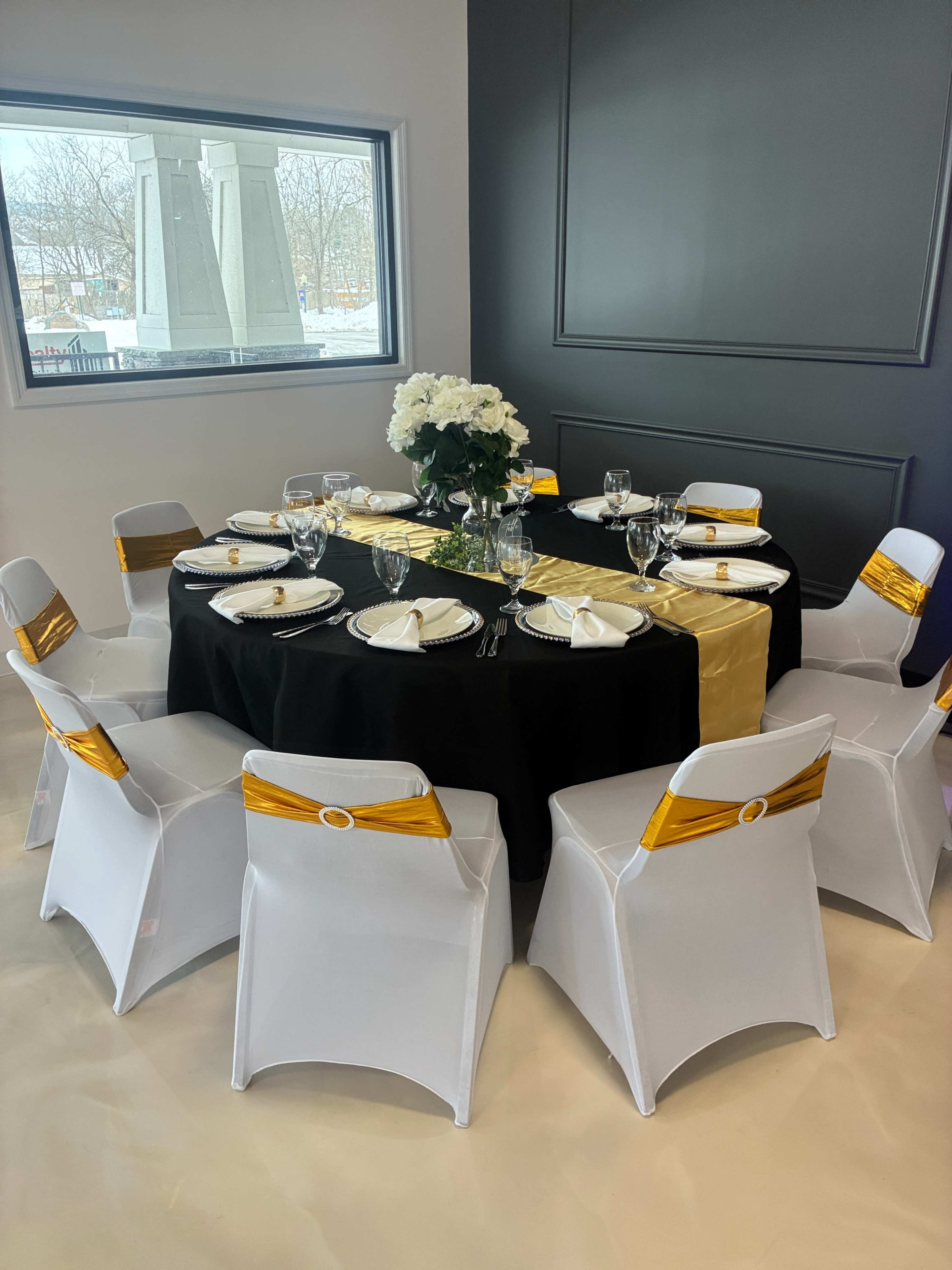 A round dining table is set with black and gold linens, surrounded by white chairs adorned with gold sashes, and topped with a floral centerpiece.