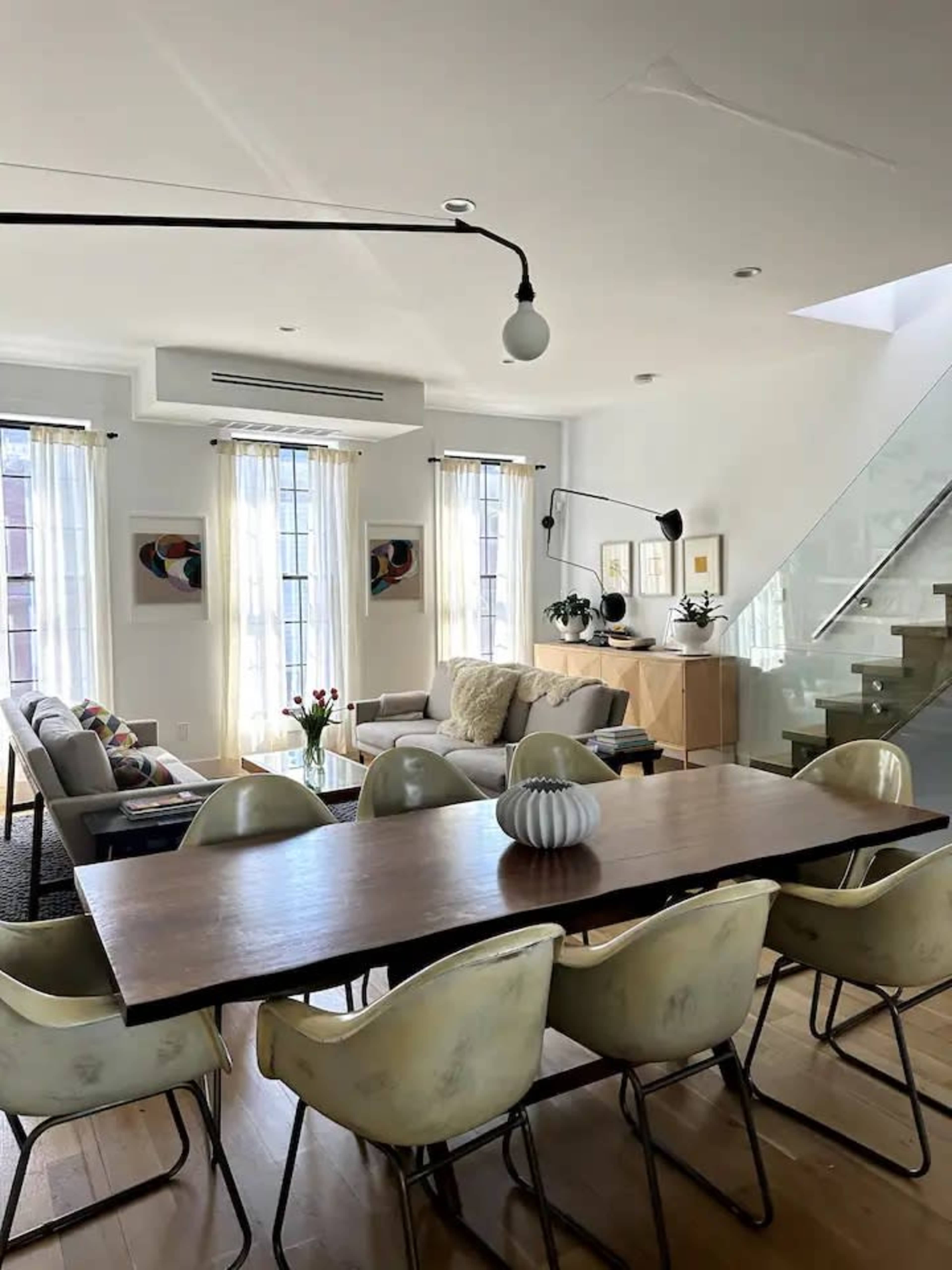 A modern living room features a large sofa, a wooden dining table with curved chairs, and large windows with sheer curtains.