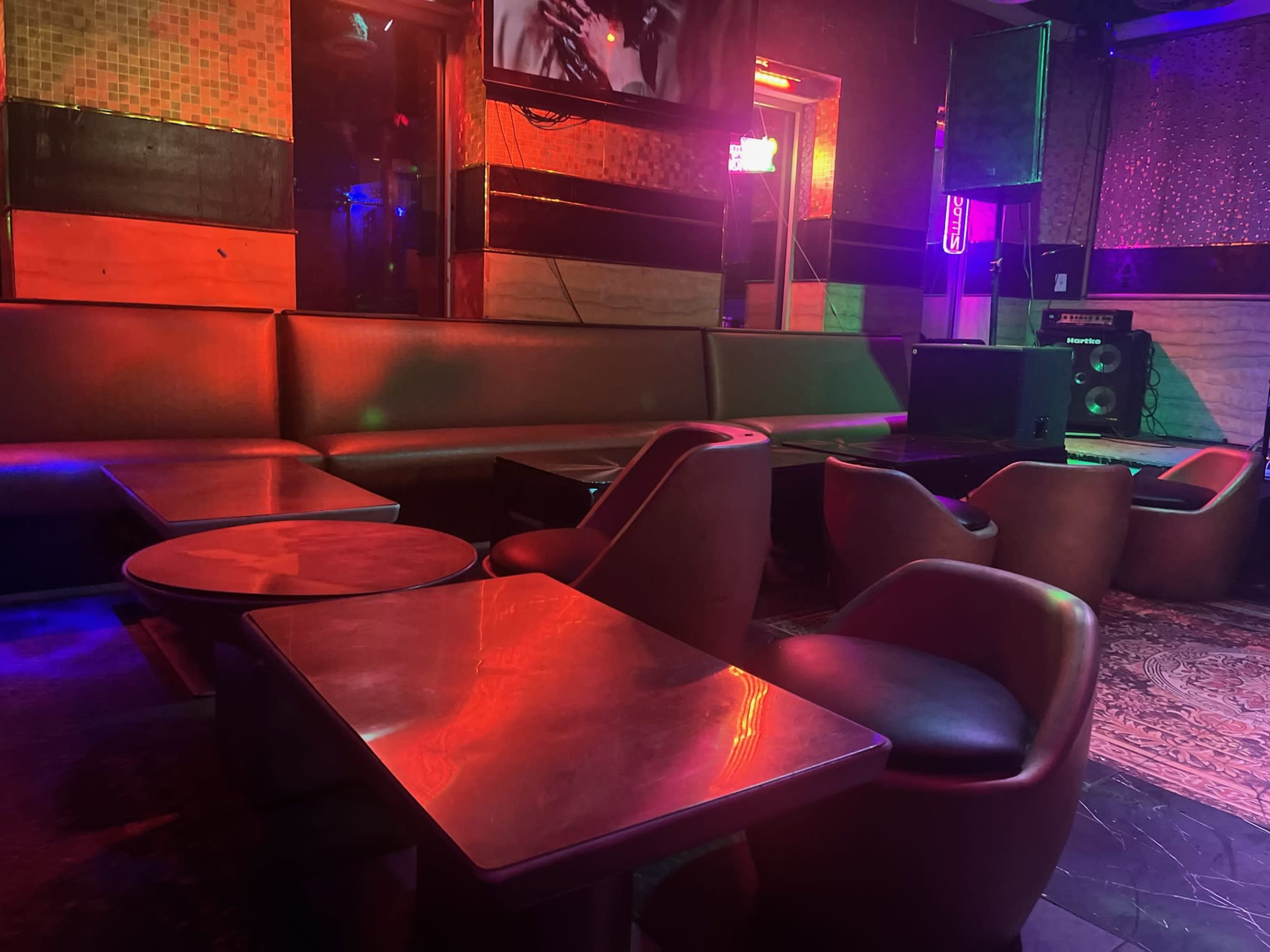 The image shows the interior of a dimly lit bar or lounge with colorful lighting, featuring several seating areas and tables.