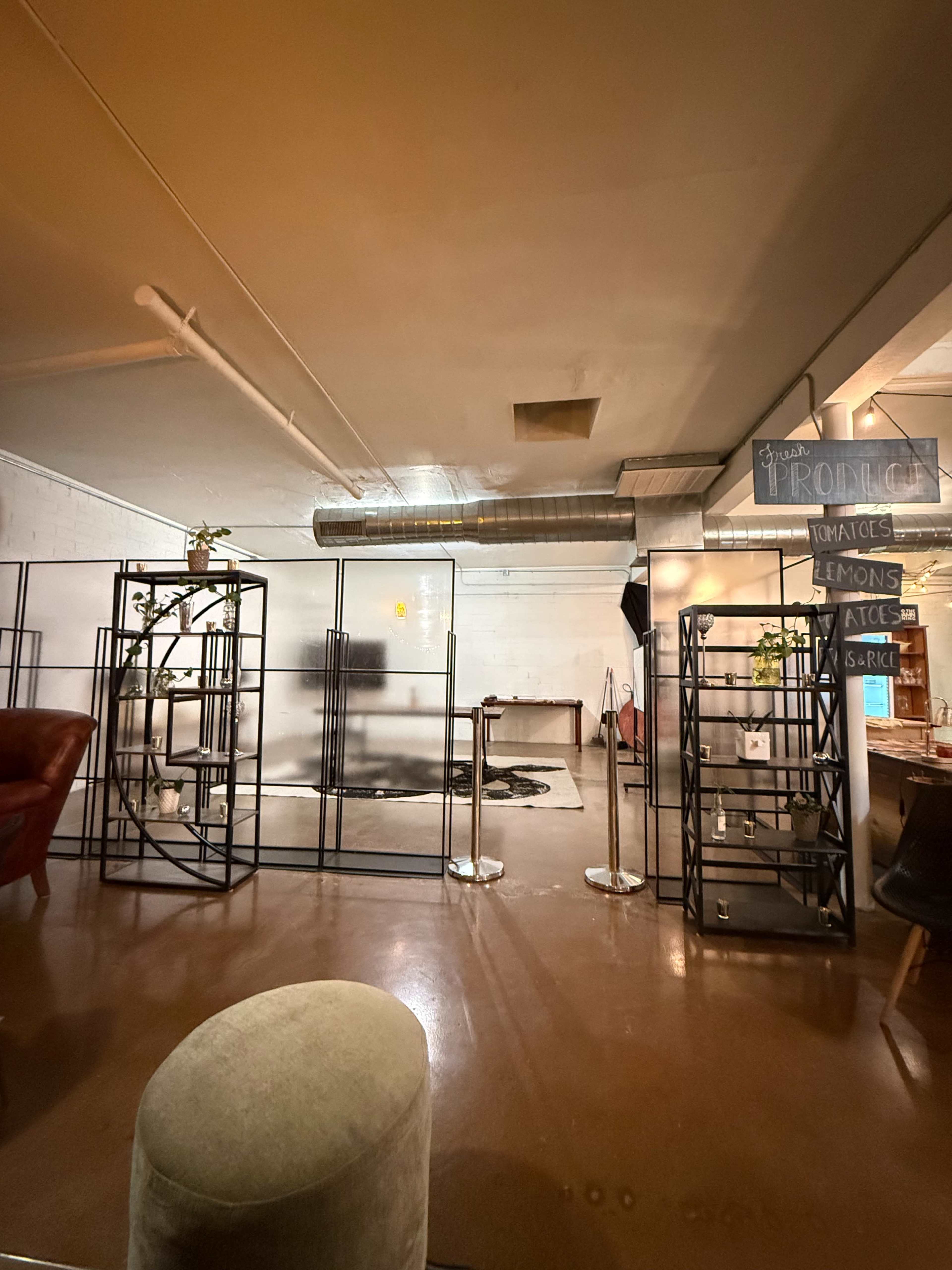 The image shows an industrial space with metal shelving units, a large ventilation duct on the ceiling, and a partially completed layout for an event or gathering area.