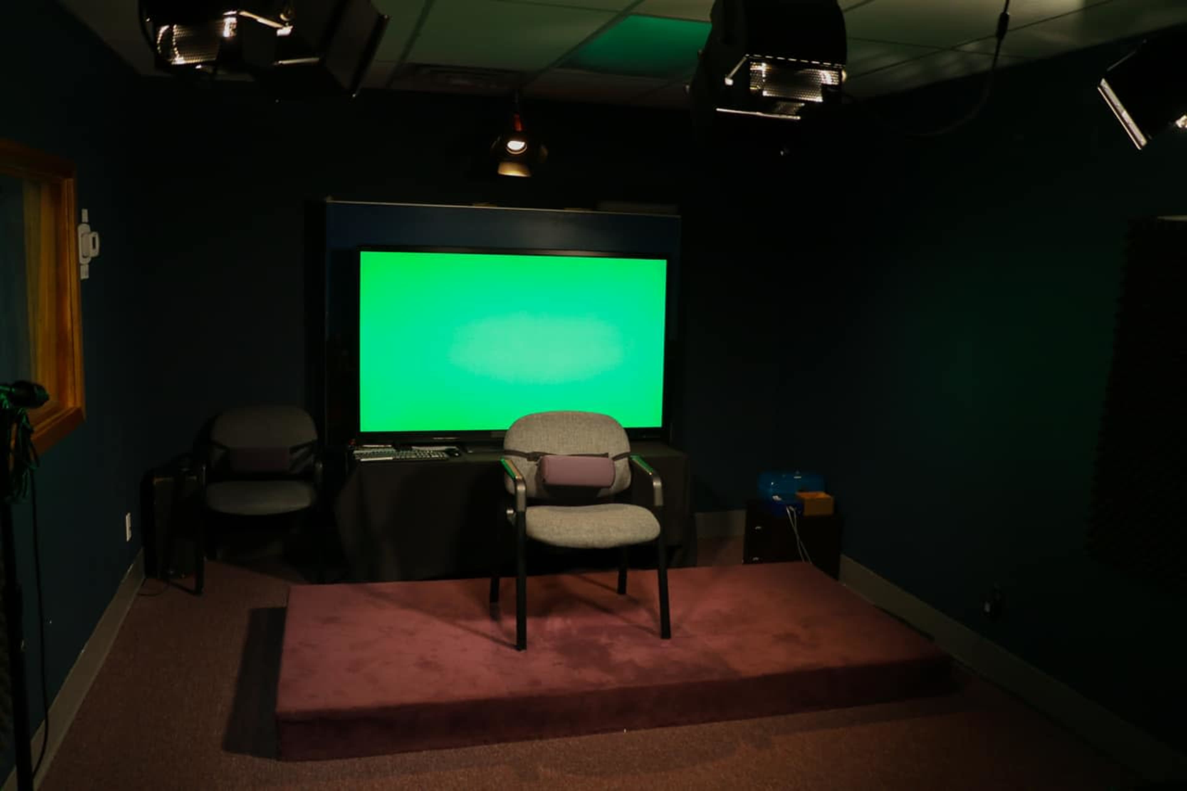 A dimly lit studio with a green screen, a chair on a raised platform, and various lighting equipment.