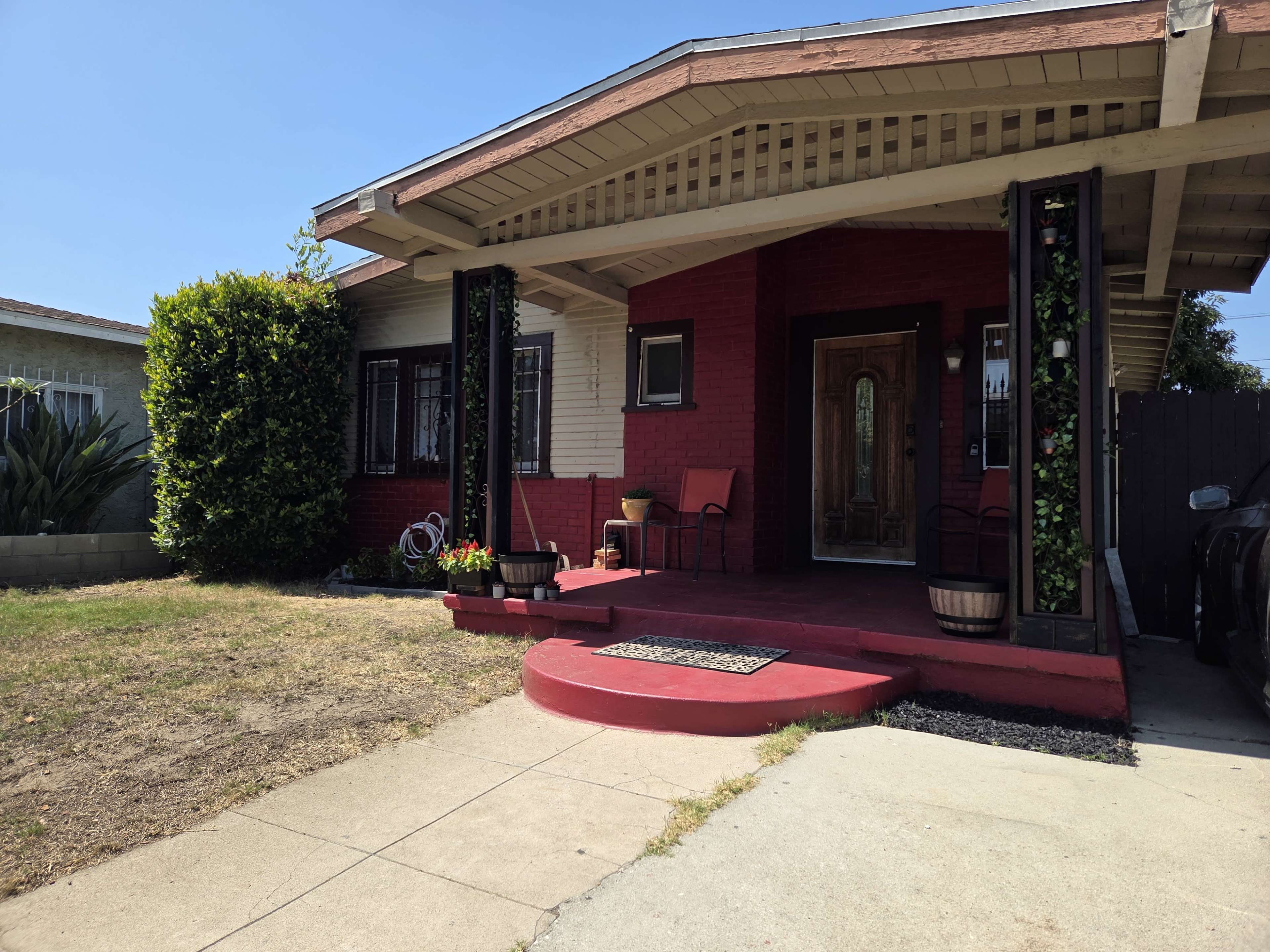 The image shows a single-story house with a red accent wall, a small front porch, and two chairs positioned near the entrance.