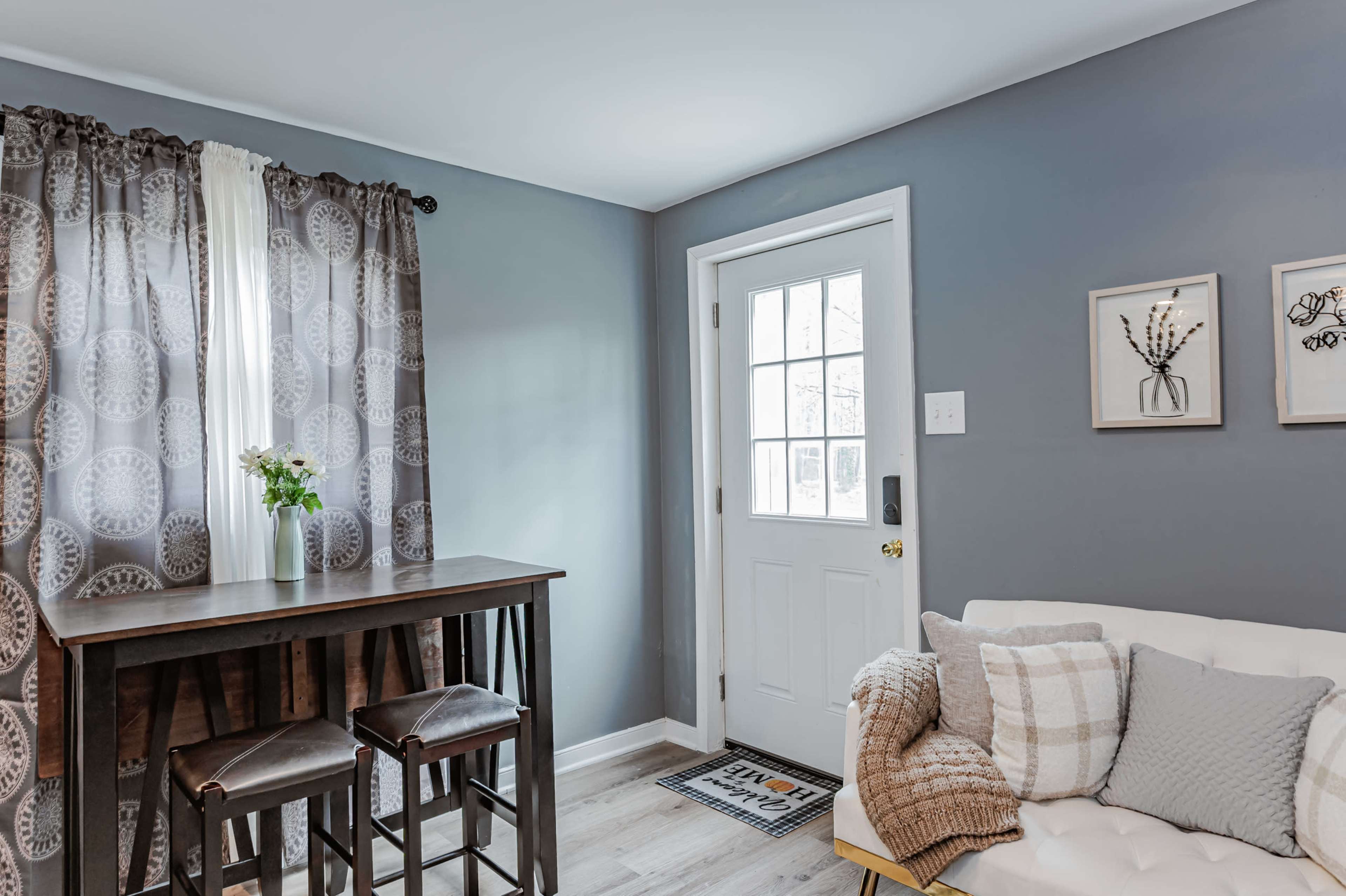 A small, modern entryway features a table with two stools, a white sofa with decorative pillows, and a door that leads outside.