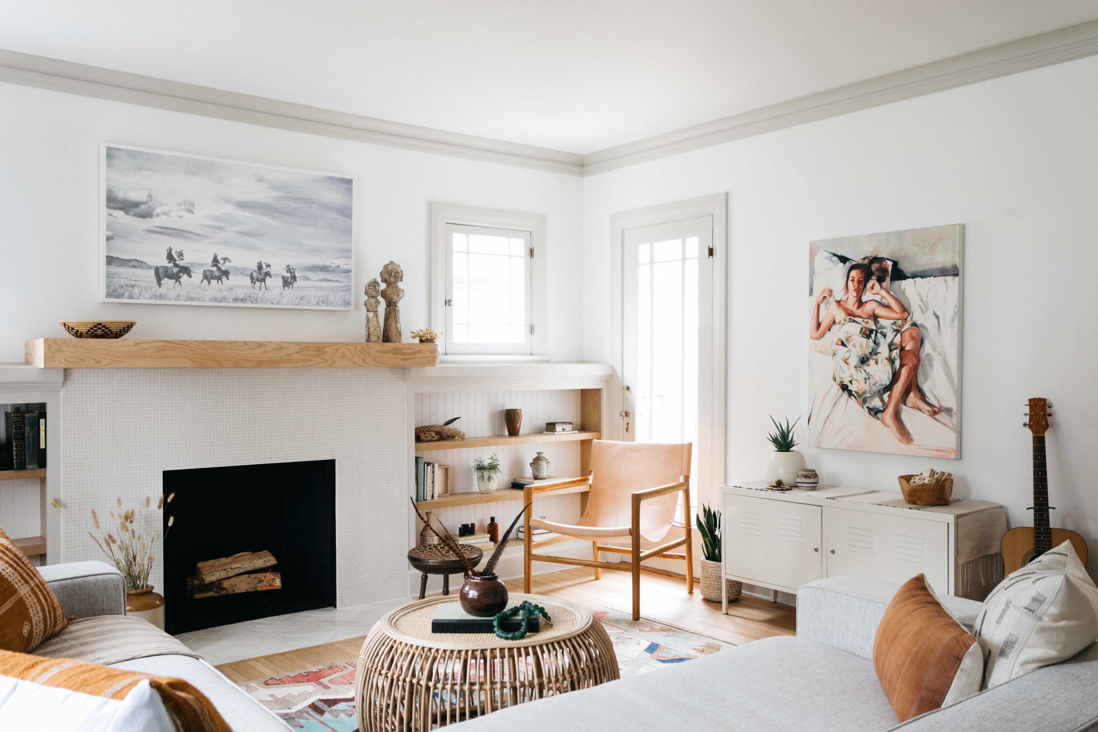 A bright living room features a white sofa, a wooden fireplace mantel, a circular rattan coffee table, and various decorative elements including plants and artwork on the walls.