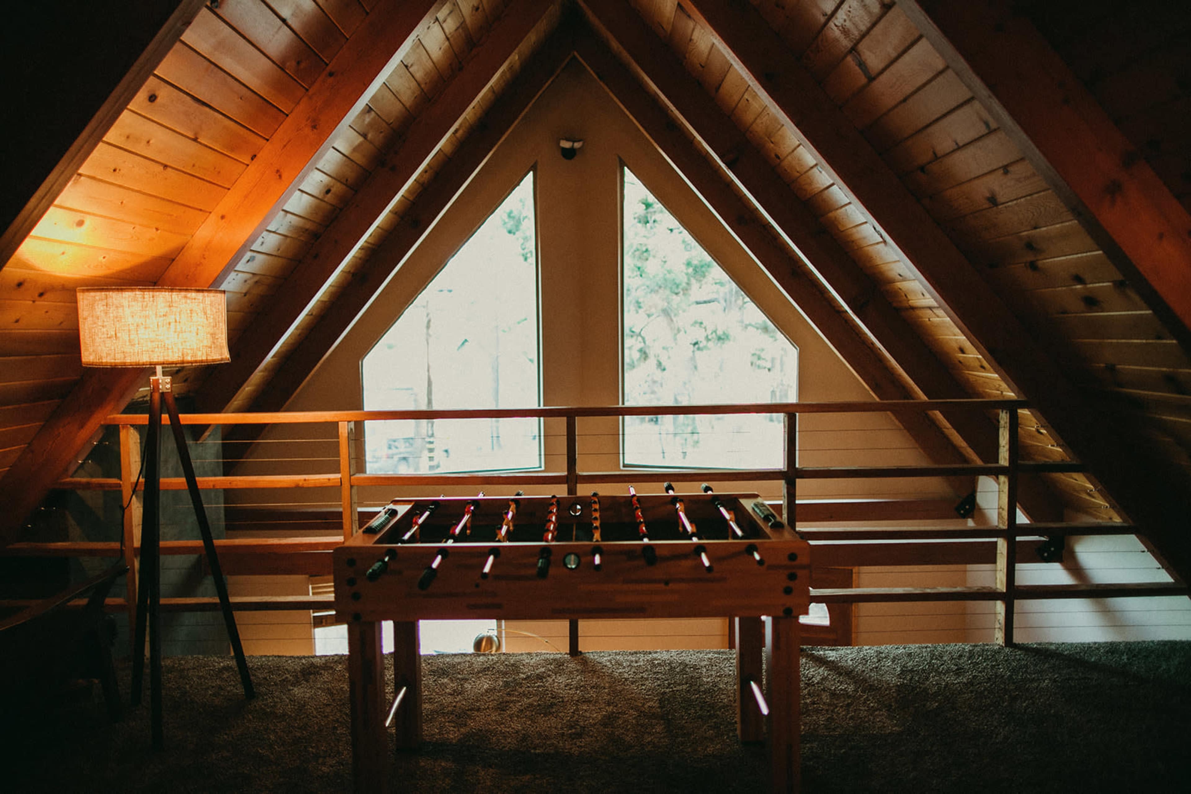 A wooden foosball table located in an attic with triangular windows and a lamp beside it.