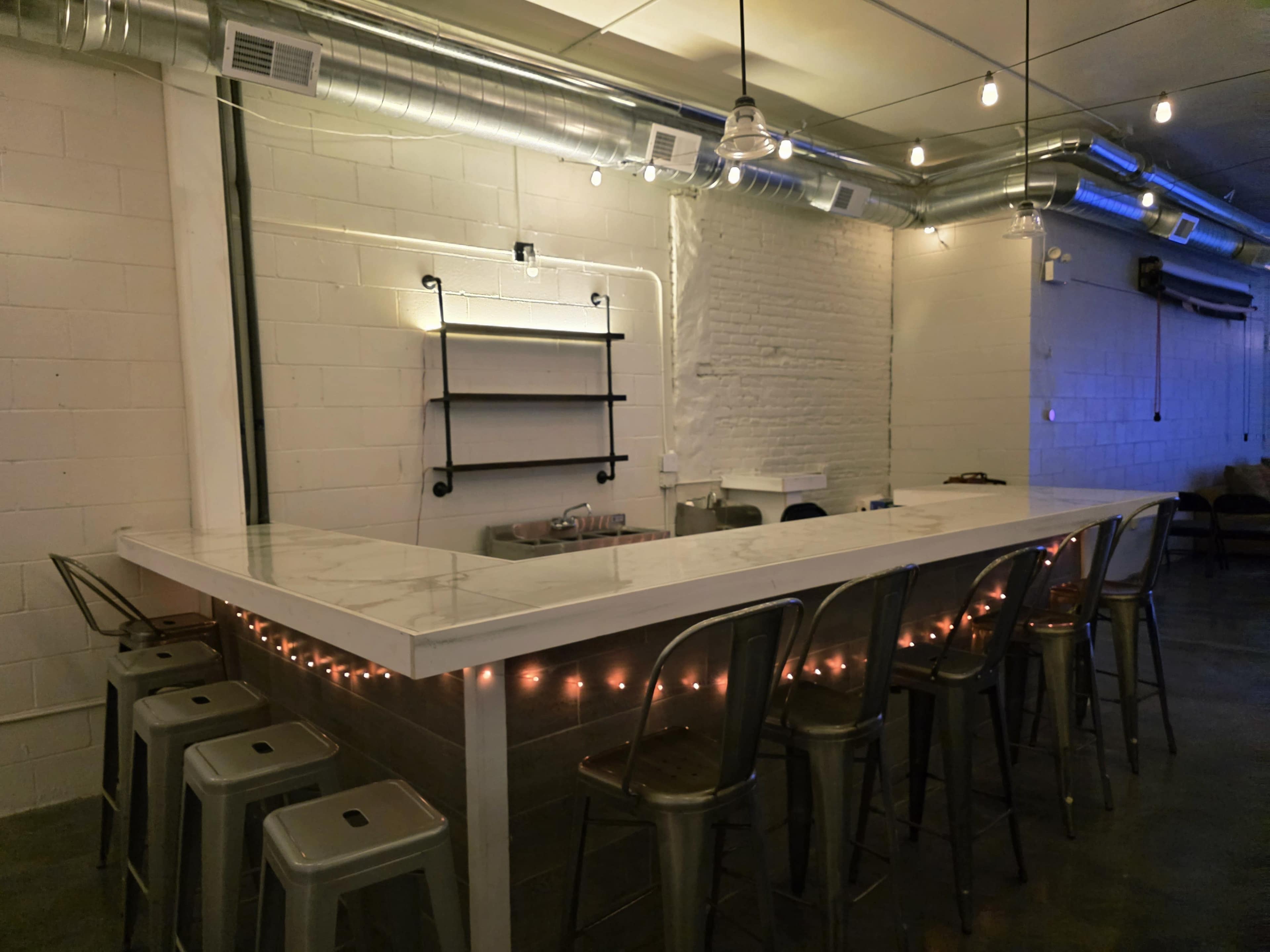 The image shows a modern bar area with a white countertop and metal stools, featuring exposed brick walls and overhead lighting.