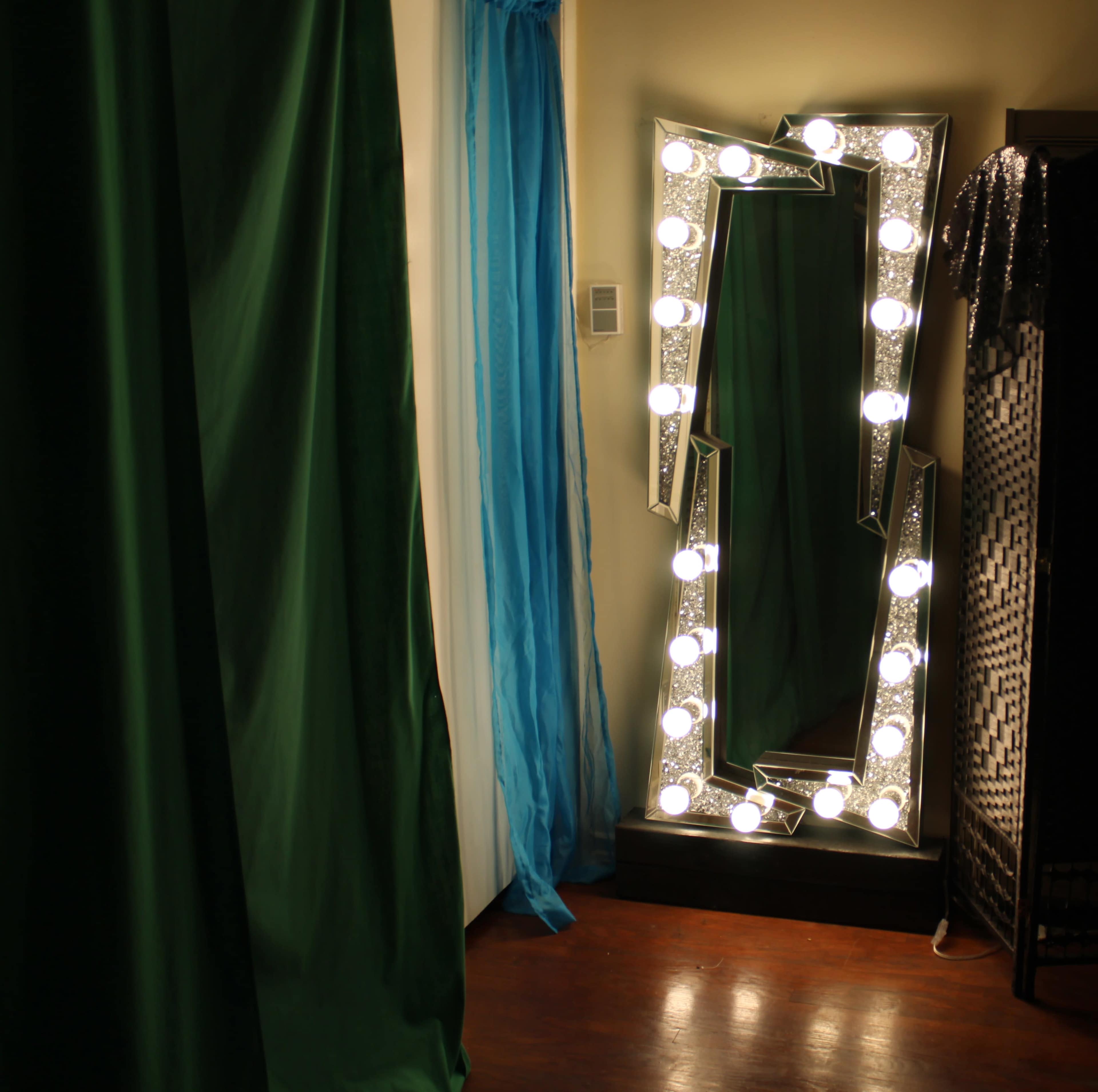 The image shows a brightly lit vanity mirror framed with decorative lights, positioned next to green curtains and a blue drape in a dimly lit room.