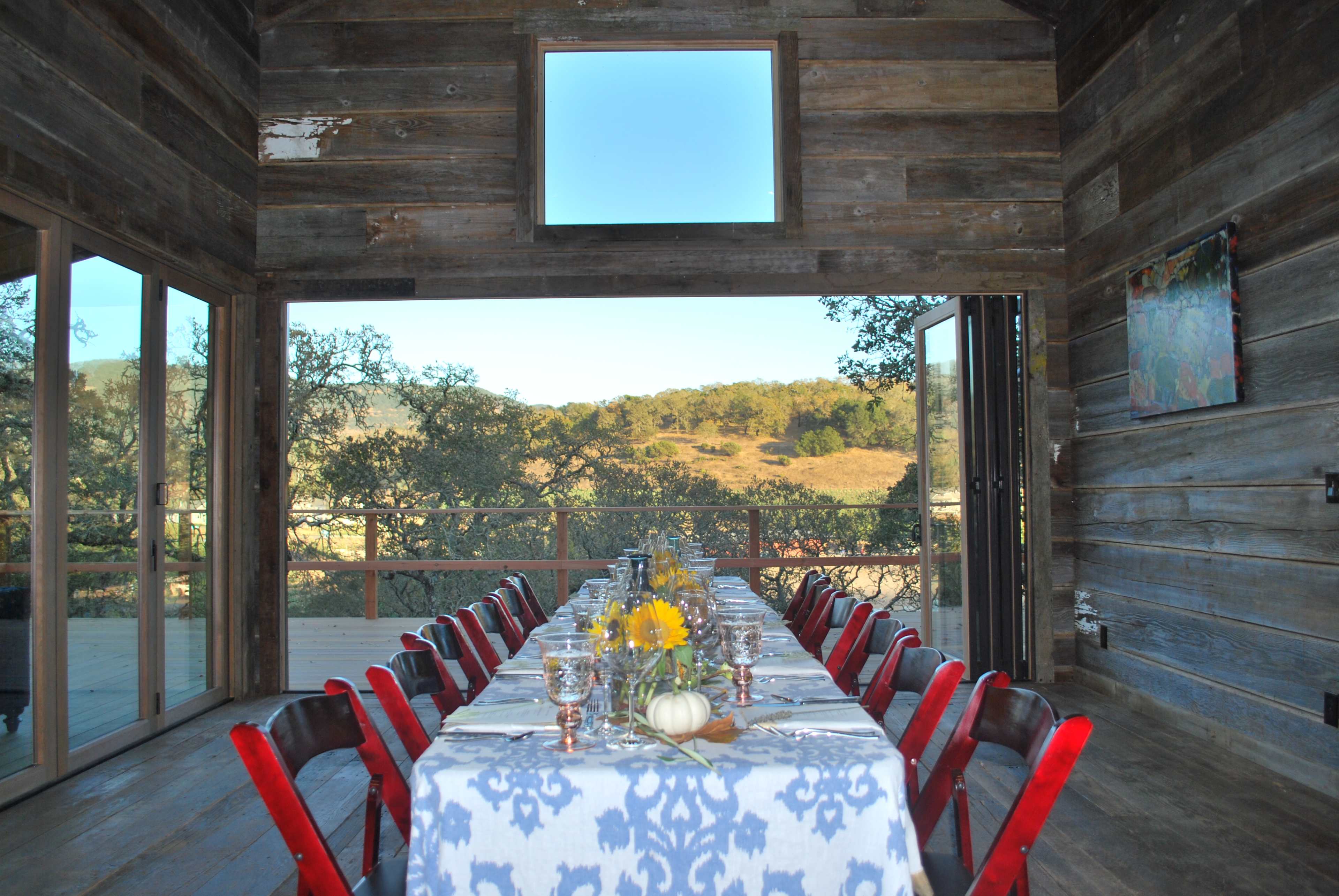 A large dining table is set with a blue and white tablecloth inside a rustic wooden room, overlooking a scenic outdoor landscape with rolling hills and trees.
