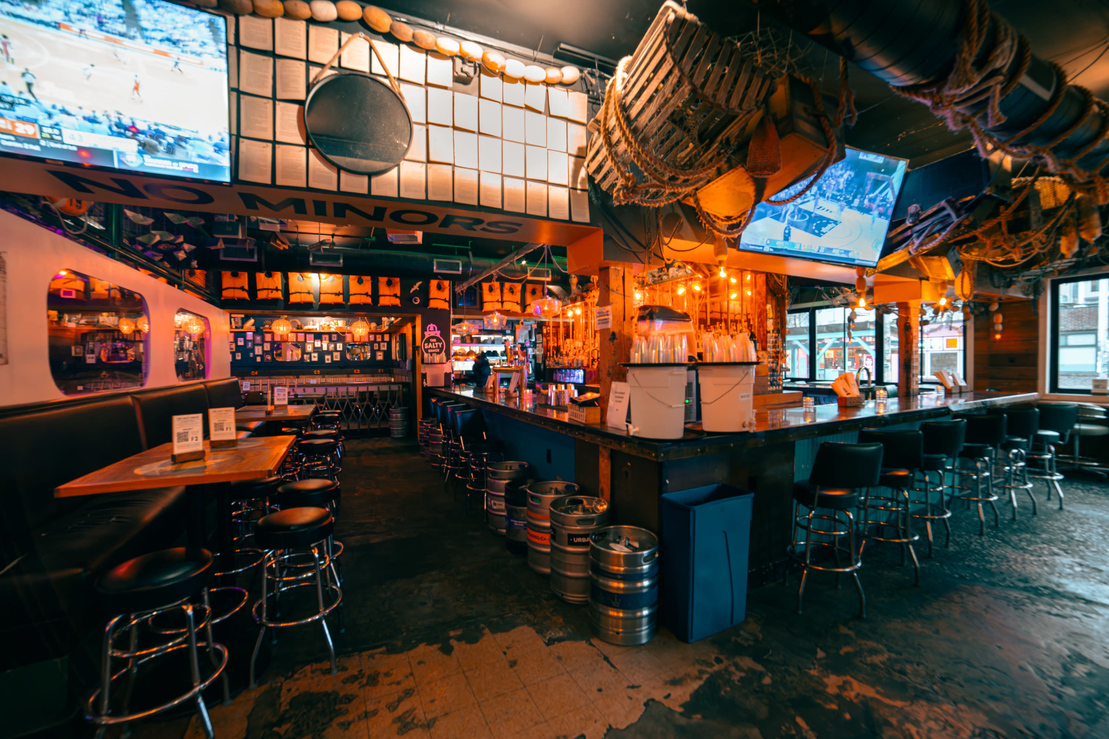 The image shows a rustic bar interior with a long counter, high stools, and multiple TVs displaying sports, illuminated by warm lighting and decorated with fishing gear.